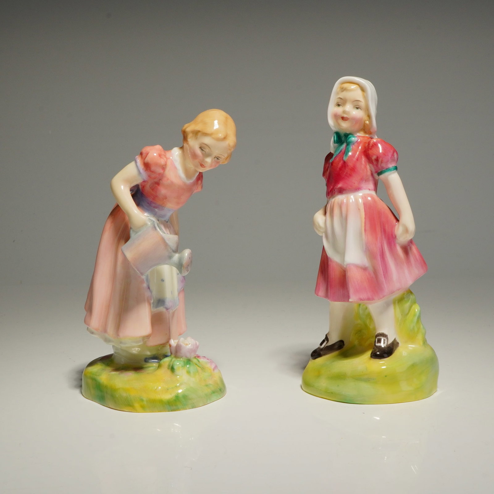 A Pair of Royal Doulton Figurines, Mary Mary HN2044 and Jill HN2061: A pair of Royal Doulton bone china figurines, Mary Mary HN2044 and Jill HN2061. Mary Mary is depicted in a pink dress with a prominent blue bow, leaning forward to water a pink blossom from a small wa