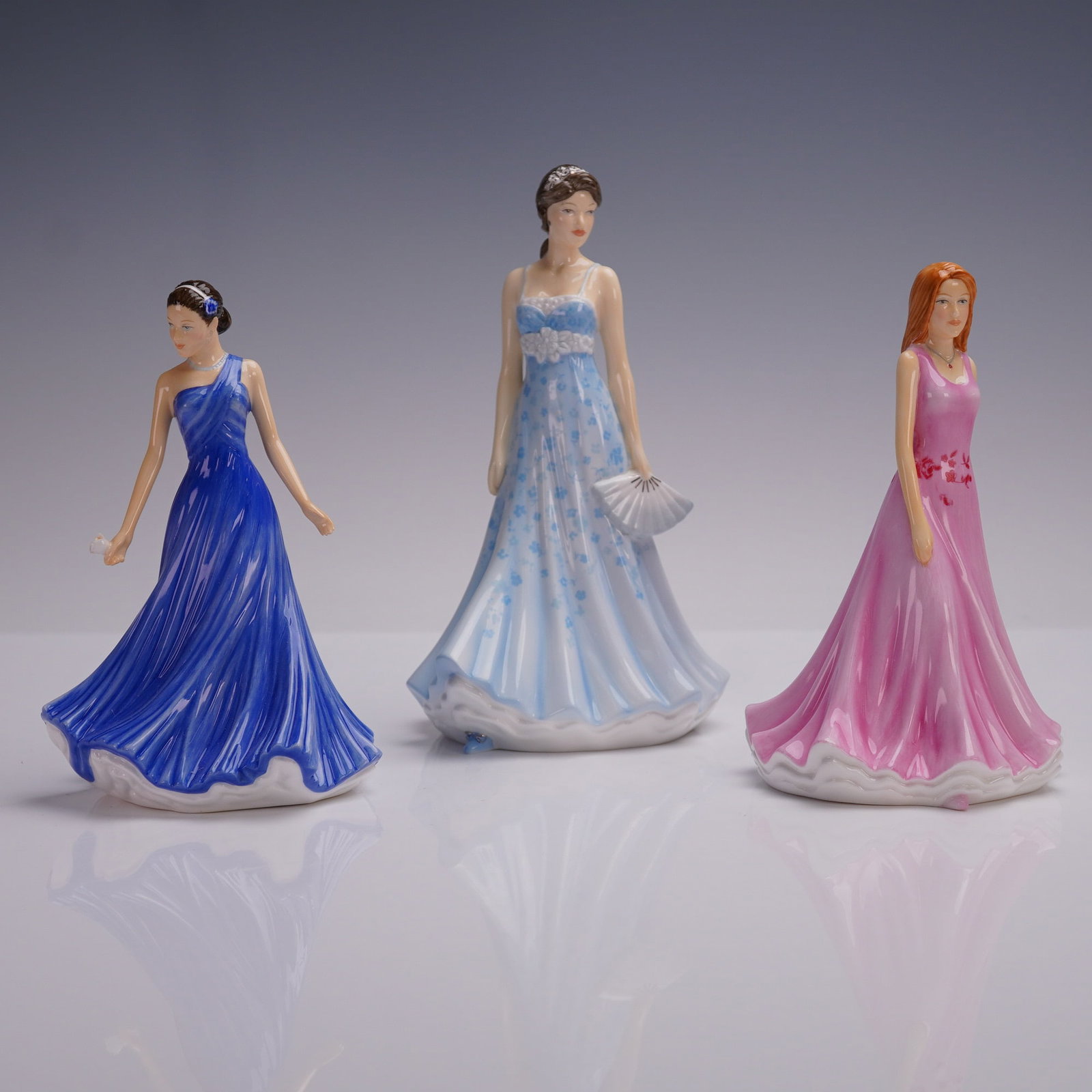 3pc Royal Doulton Figurines Set, Lottie, Stephanie, Liz: This lot features three bone china figurines. The set includes: Lottie HN5776, A young woman in a deep blue, one-shoulder gown adorned with a flower in her hair and holding a small object in her hand.