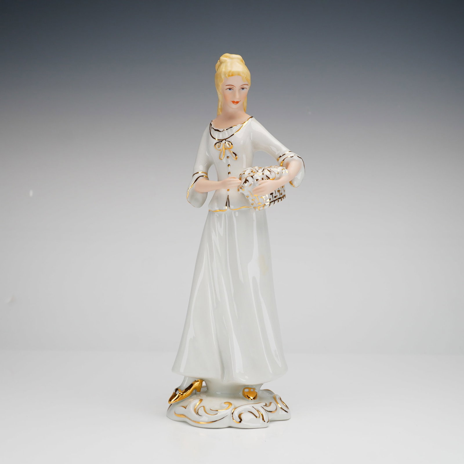 Royal Dux Bohemia Porcelain Figurine, Woman in White: This Royal Dux Bohemia porcelain figurine of a woman holding a basket is finished in a white glaze with gilt accents. The figure is modeled in a stylized 18th-century gown and stands upon a scrolled b