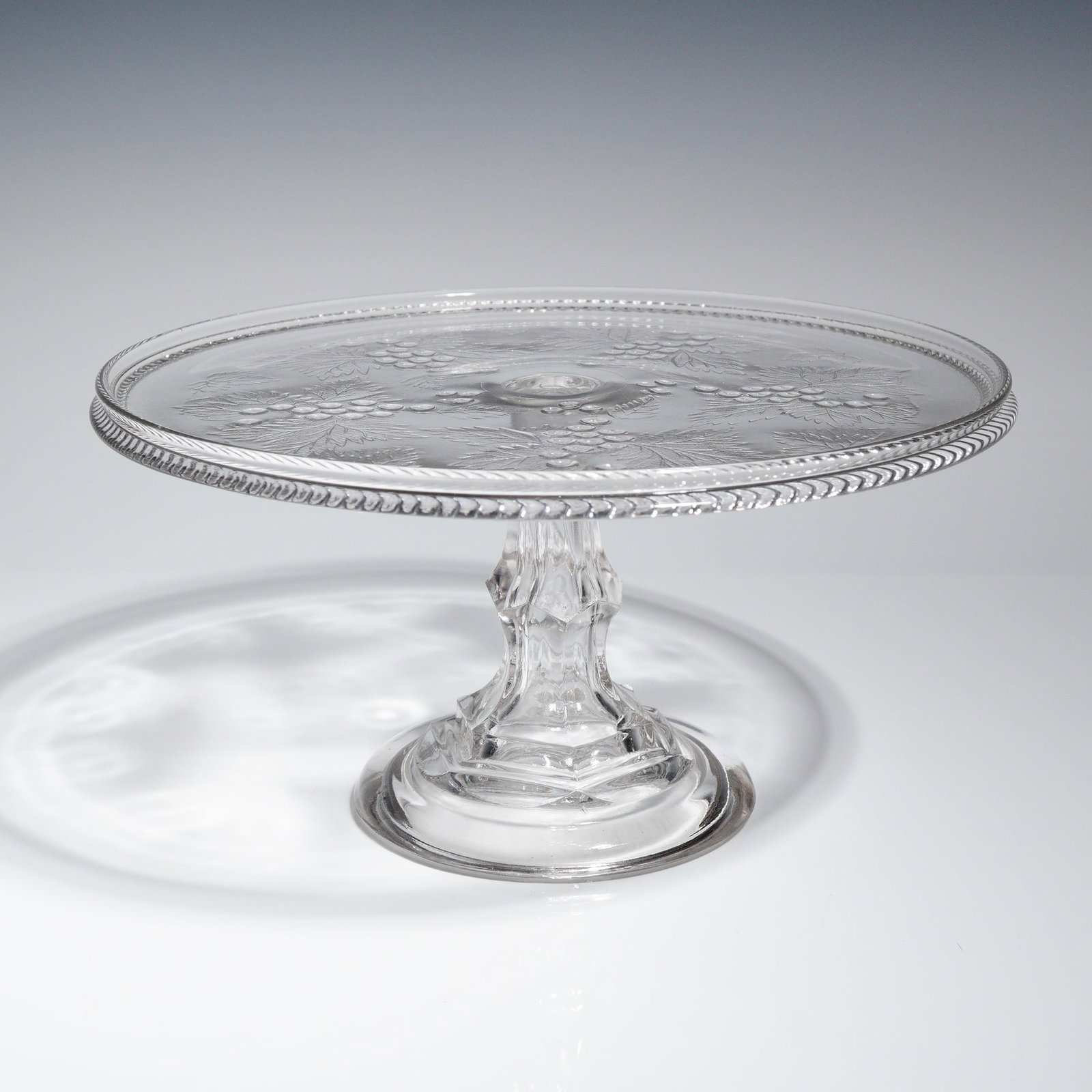 Adams & Co. EAPG Good Luck Pattern Pressed Glass Pedestal Cake Stand: A single Early American Pressed Glass (EAPG) pedestal cake stand manufactured by Adams & Co. in the 'Good Luck' pattern, also known as 'Leaf & Berry'. The object is constructed from clear, colorless p