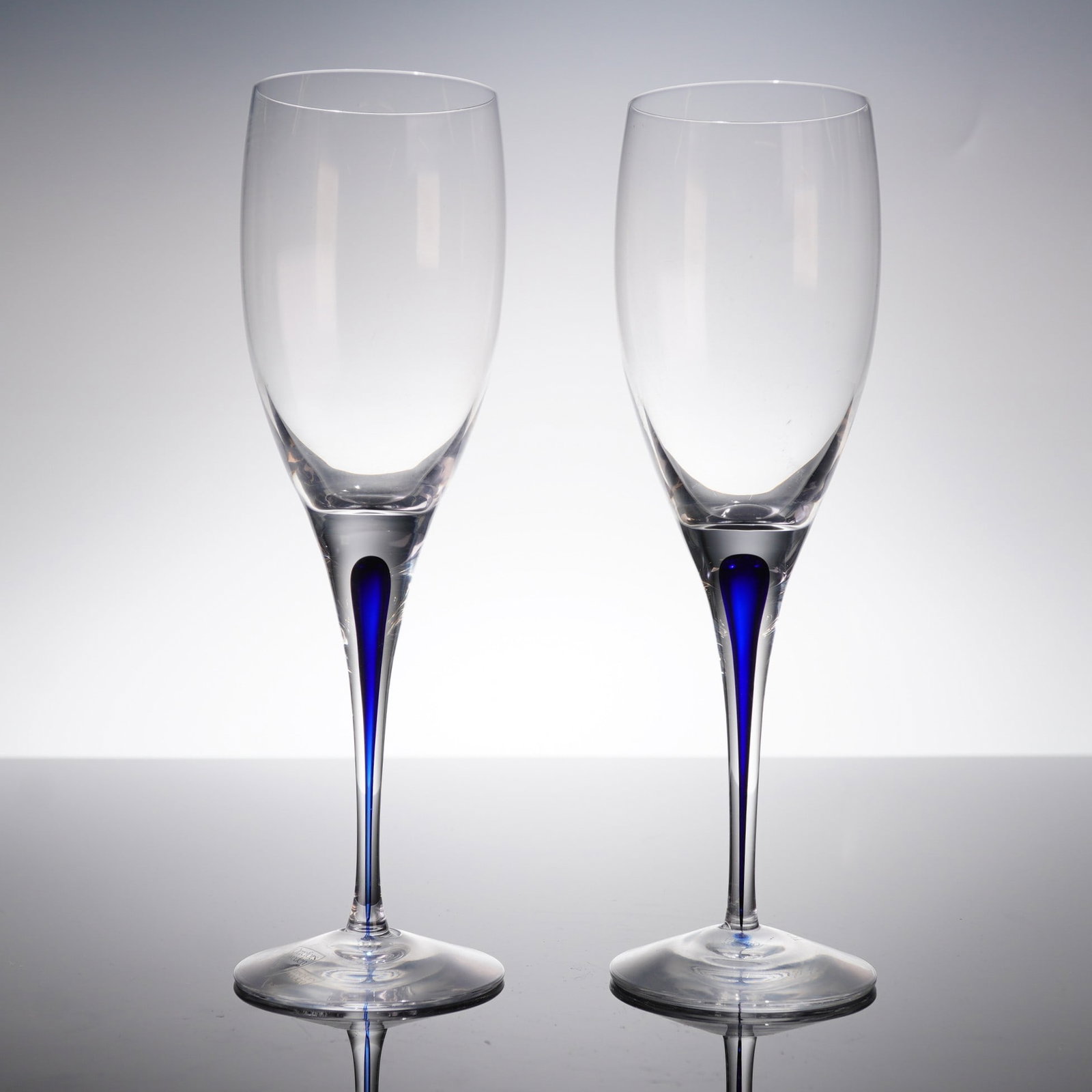 A Pair of Orrefors Intermezzo Blue Crystal Wine Glasses: A pair of crystal stemware glasses from the Orrefors Intermezzo Blue line. The design features an elongated, clear bowl suitable for white wine service, supported by a tall, slender stem. The pieces e