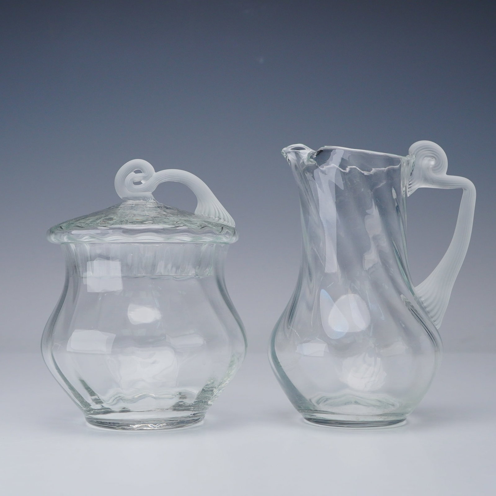 2pc Clear and Frosted Art Glass Creamer and Lidded Sugar Set: This clear art glass set includes a hand-blown creamer with a ribbed applied handle and shaped spout, paired with a matching sugar vessel and fitted lid. The creamer features a fluid, slightly twisted