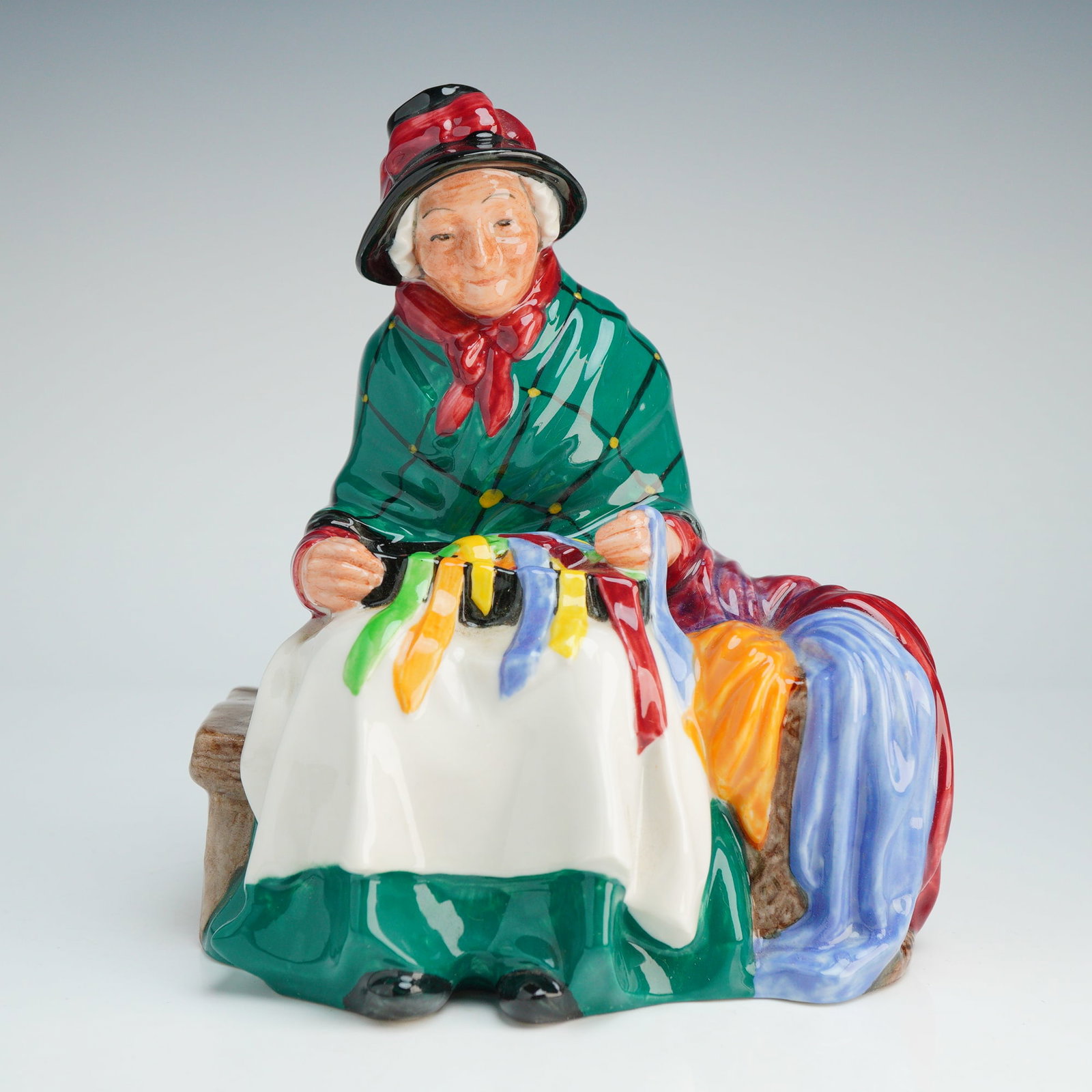 Royal Doulton Figurine, Silks and Ribbons HN2017: The figure depicts an elderly female street vendor seated on a brown wooden bench. She is wearing a green plaid shawl over a white apron and green skirt, with a red neckerchief and a black bonnet trim