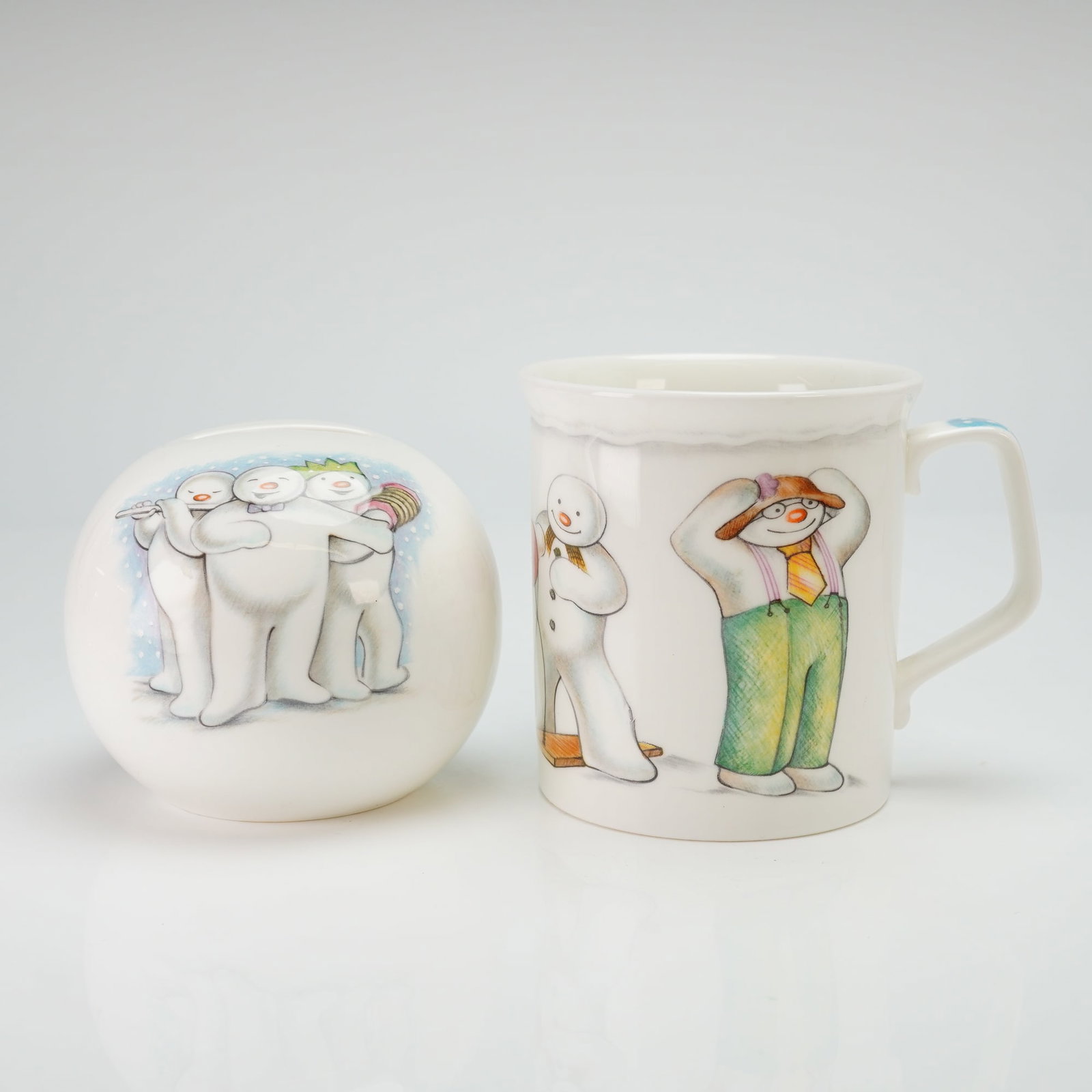 2pc Royal Doulton The Snowman Gift Collection, Mug and Coin Bank: A 2pc Royal Doulton bone china set from The Snowman Gift Collection, dating to 1985. The set includes a spherical Snowball money bank with a recessed coin slot at the top and a handled mug titled Play