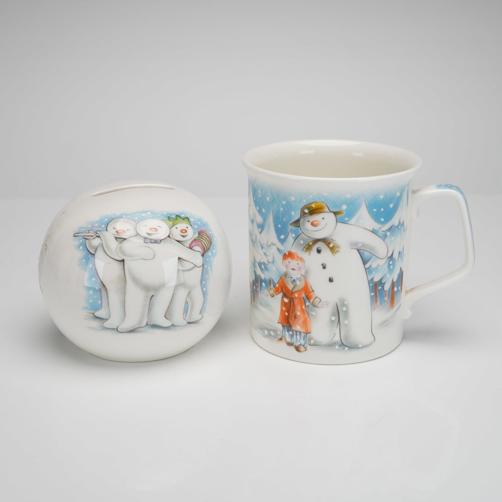 2pc Royal Doulton The Snowman Gift Collection Mug & Money Bank: Two Royal Doulton bone china items from The Snowman Gift Collection, featuring illustrations based on the characters by Raymond Briggs. The lot includes a spherical Snowball money bank and a mug title
