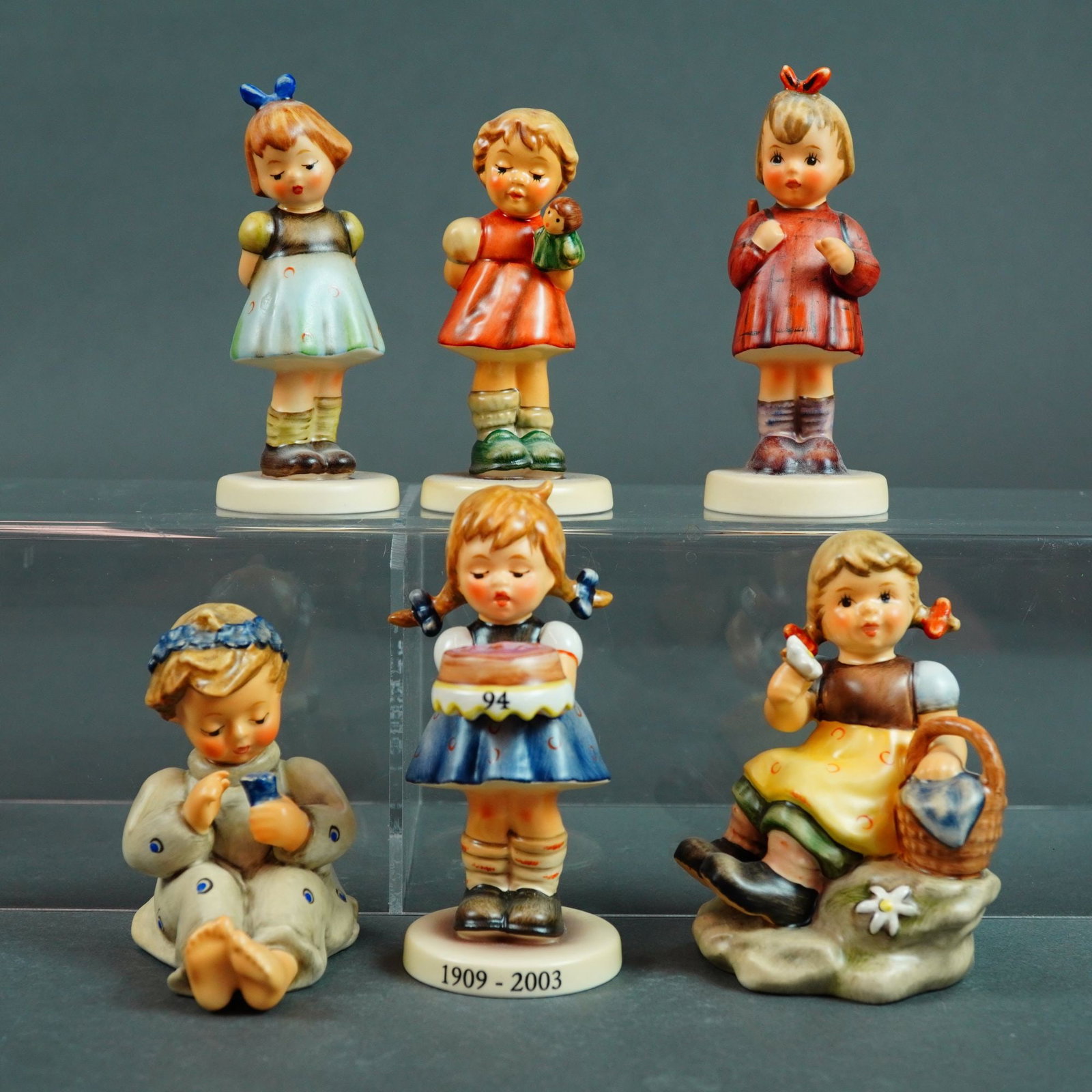 Goebel M.I. Hummel, Collection of Six Girl Figurines: This lot features a collection of six (6) hand-painted Goebel M.I. Hummel ceramic figurines, primarily featuring young girl subjects. Each piece is crafted from slip-cast porcelain with a signature ma