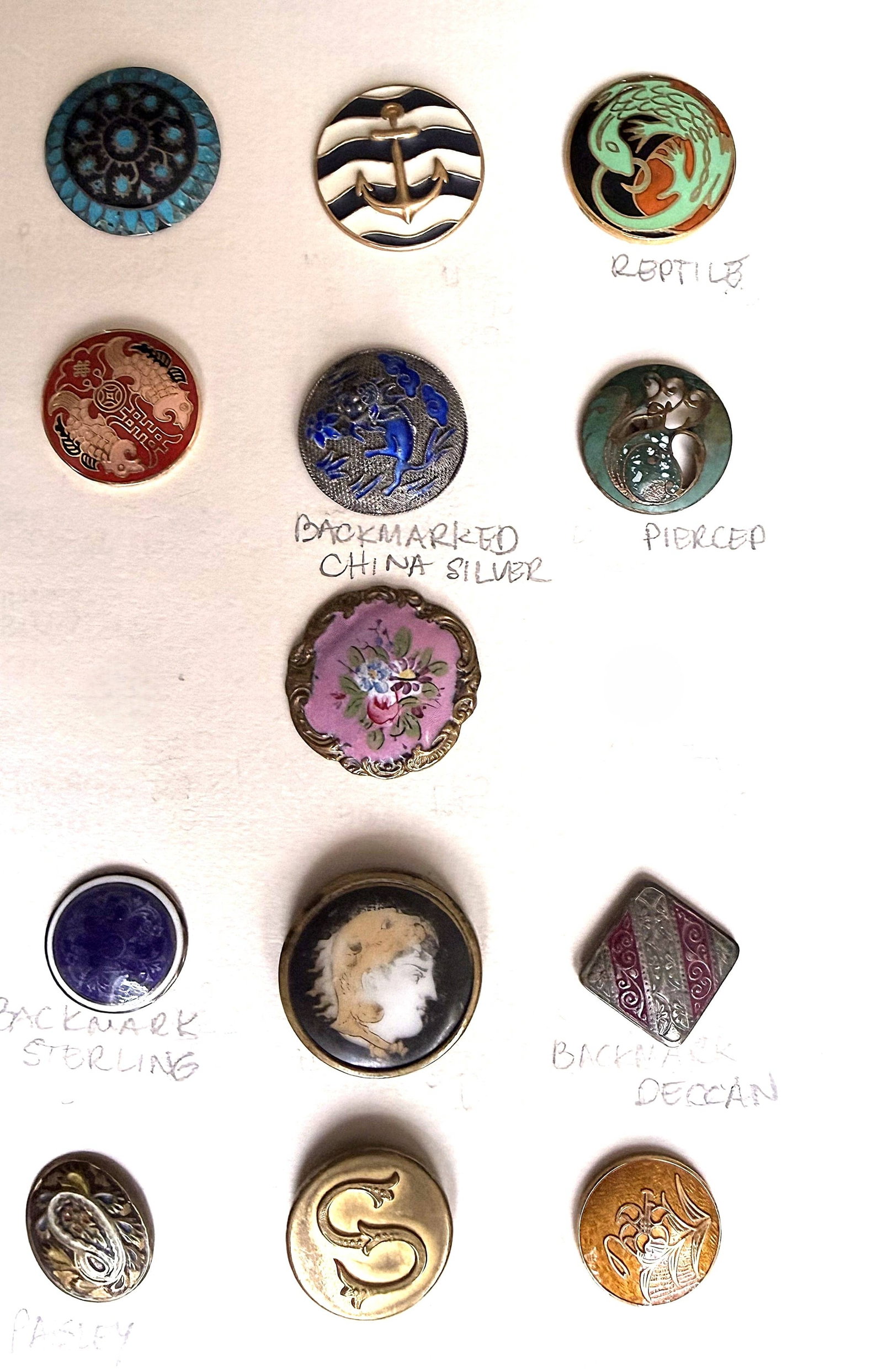 A SMALL CARD OF ASSORTED ENAMEL AND PORCELAIN BUTTONS: 13 Medium 19th/20th C. Assorted technique examples. This card includes a nice 19th Century silver and enamel backmarked from China cool Foo Lion, a square enamel on silver Deccan examel, a Liverpool t