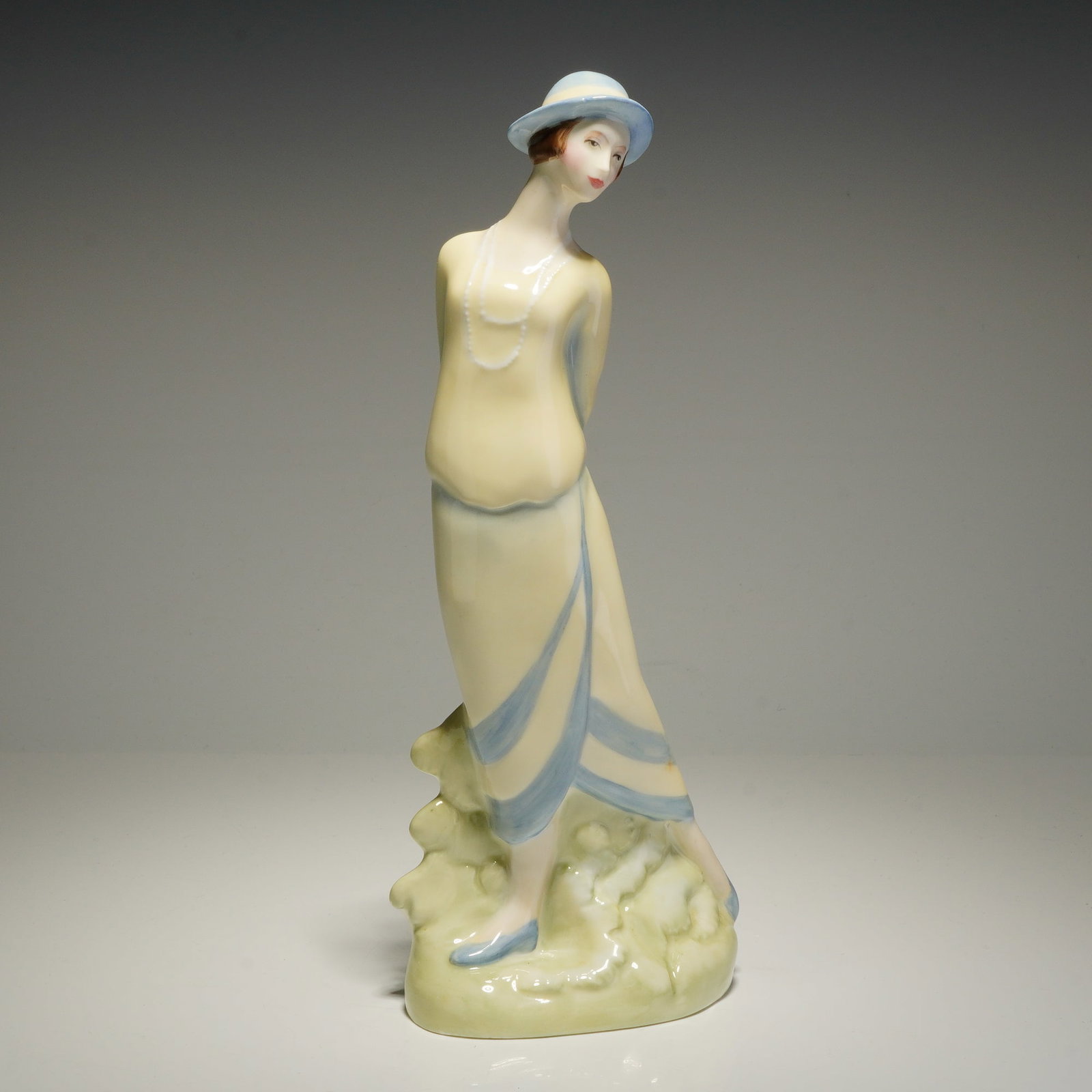 Royal Doulton Figurine, Eliza HN3799: This Royal Doulton bone china figurine, titled Eliza (HN 3799), is modeled in a tilted pose wearing a long, pale yellow dress with blue-banded trim, a matching blue hat, and blue shoes, accented by a