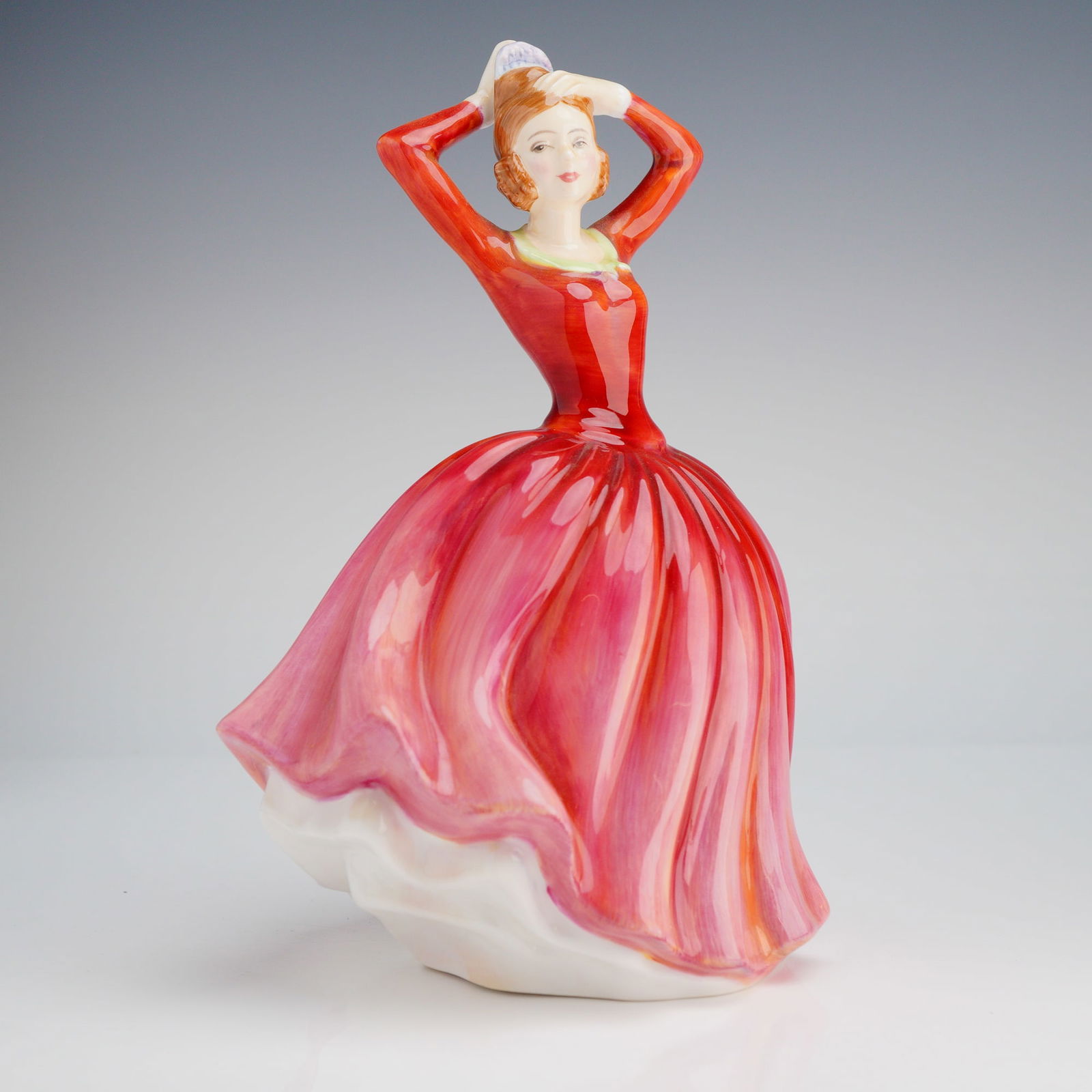 Royal Doulton Figurine, Katrina HN2327: The figure depicts a young woman in a dynamic pose with her hands raised to her head, dressed in a voluminous, flowing gown decorated in shaded tones of deep red and pink over a white petticoat. The p