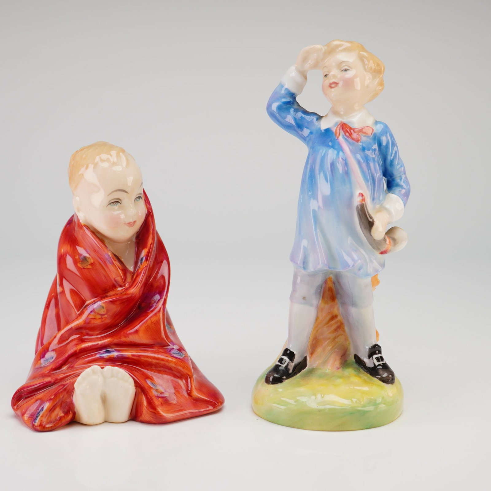 Royal Doulton Figurines, This Little Pig HN1793 and Little Boy Blue HN2062: A pair of Royal Doulton glazed bone china figurines depicting classic nursery rhyme characters. The first, titled This Little Pig (HN 1793), features a seated infant wrapped in a vibrant red patterned
