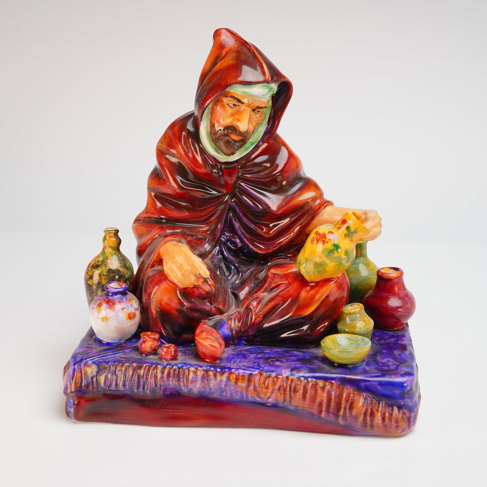 Royal Doulton The Potter Figurine HN1493: This Royal Doulton bone china figurine, titled The Potter (model HN 1493), depicts a seated merchant in Middle Eastern-style attire. The figure is dressed in a deep red hooded robe and a green turban,