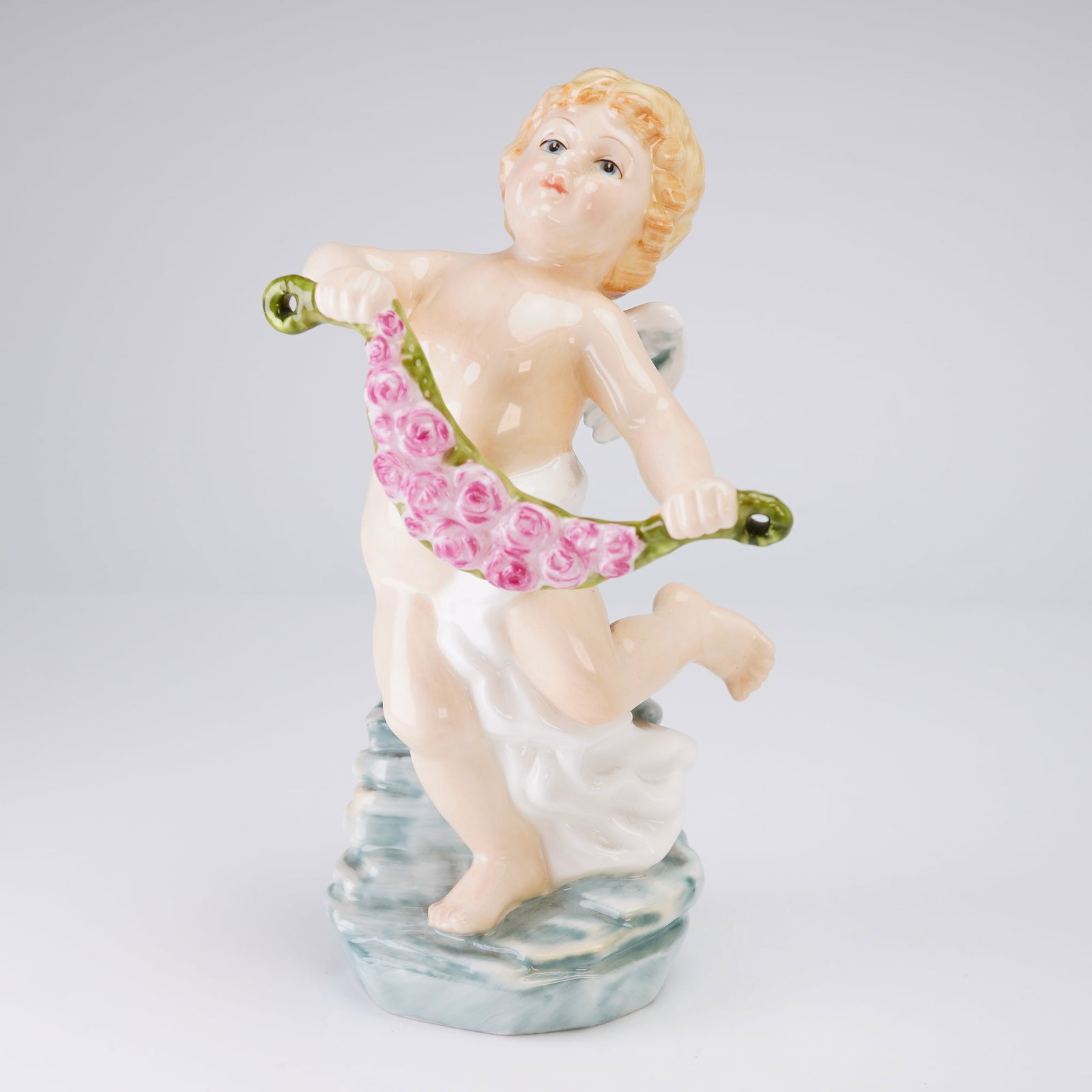 Doulton International Cherubs of the Arts Figurine, Dance: The figure depicts a putto or cherub standing in a dynamic pose on a stylized base, holding a garland of pink roses. The cherub features fair skin tones, light yellow hair, and small white wings. The