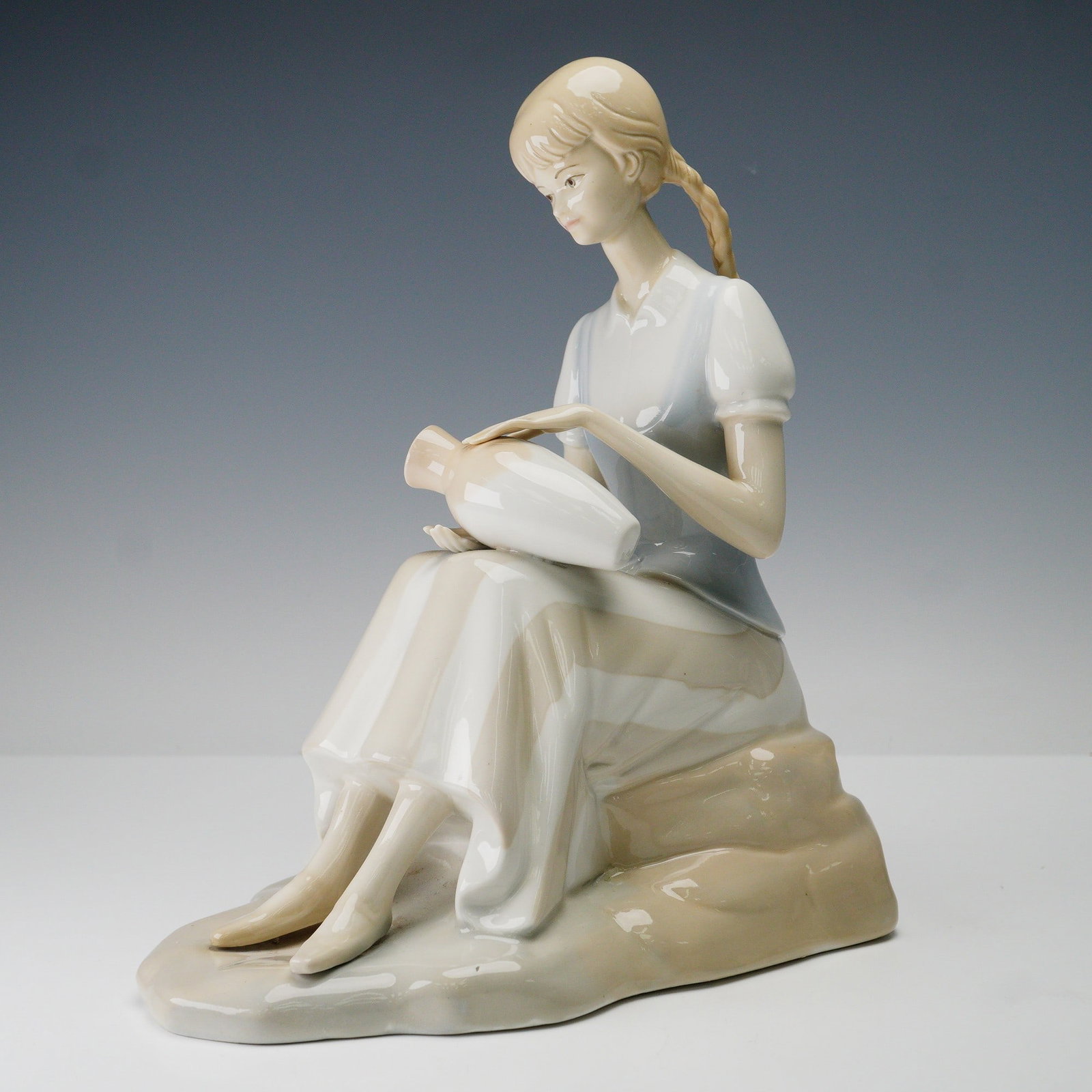 Porceval Spain Porcelain Figurine, Girl with Vase: The figure is crafted in soft pastel tones with a glossy glaze, highlighting delicate details such as the girl's braided hair, serene expression, and flowing dress. She is shown resting on a naturalis