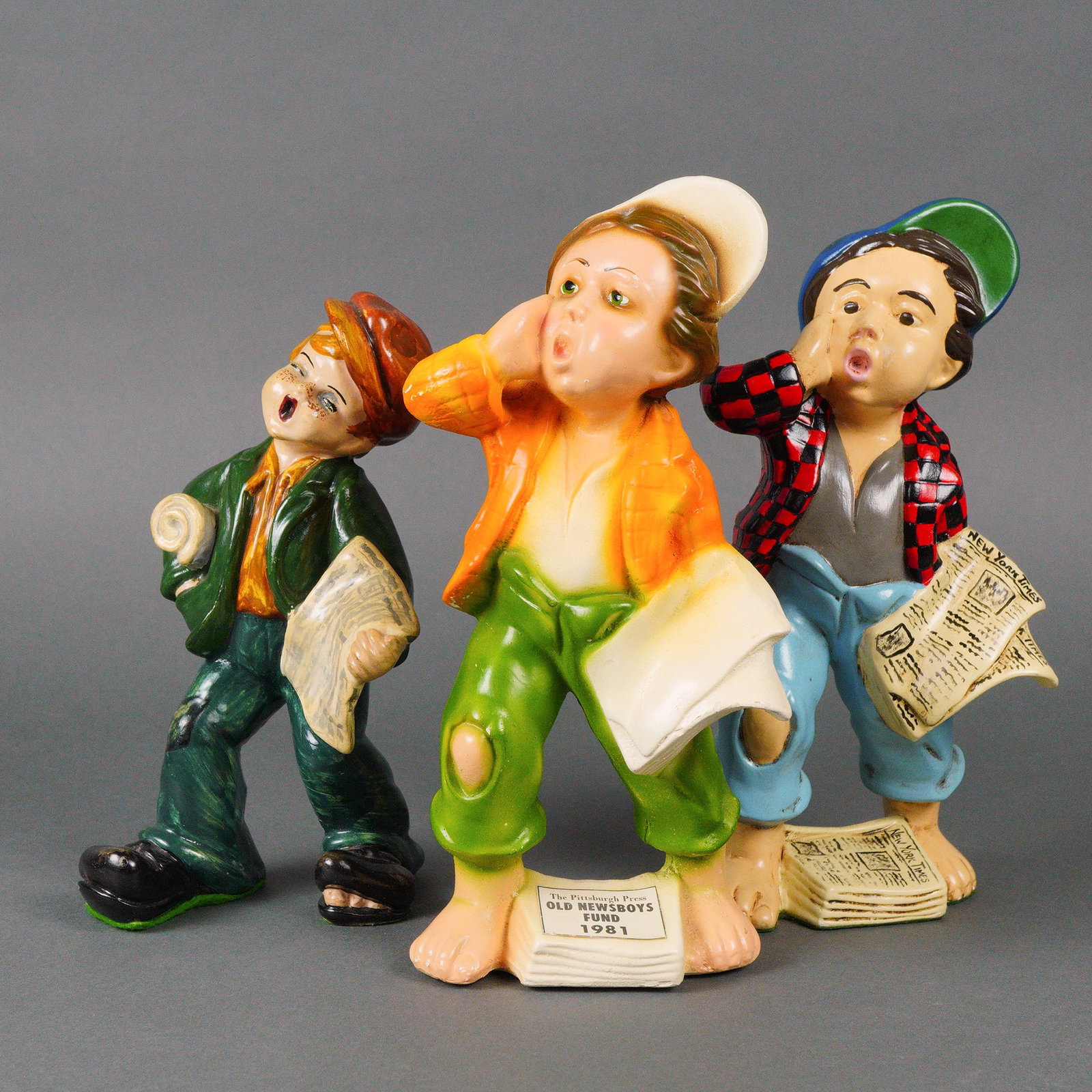 A Set of Three Newsboy Ceramic Figurines: A set of three ceramic newsboy figurines, each molded and hand painted in a bright, caricatured style with expressive faces and exaggerated poses. The figures wear colorful clothing with patched or to