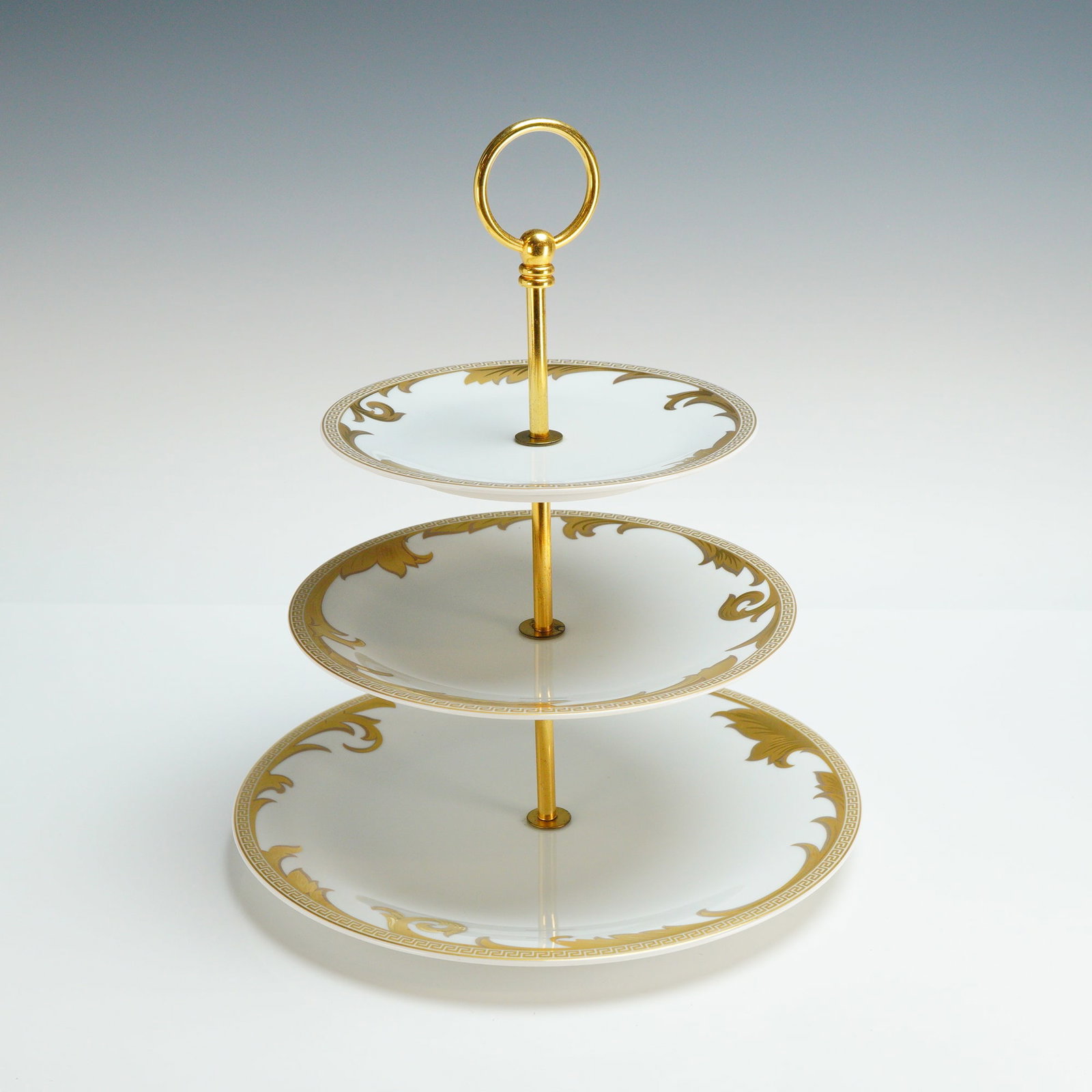 Rosenthal Versace Three-Tiered Cake Stand, Arabesque Gold: The etagere consists of three graduated circular white porcelain plates supported by a central gold-tone metal rod culminating in a circular ring handle. The plates are decorated with gold-tone foliat