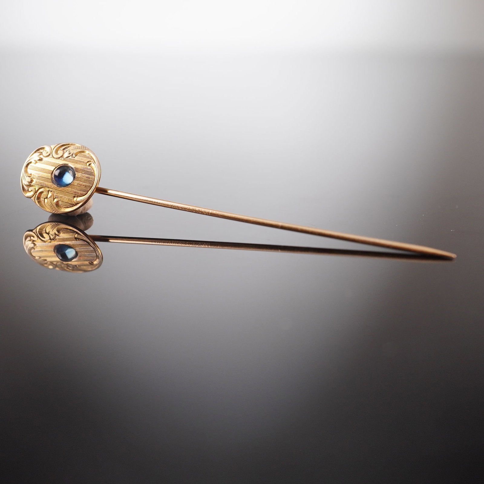 14K Gold Sapphire Engraved Scrollwork Stick Pin: A yellow gold stick pin featuring a head with engraved scrollwork detailing and set with a sapphire cabochon. The stem is marked 14K. Dimensions2.75"L x 0.5"W Weight1.8g