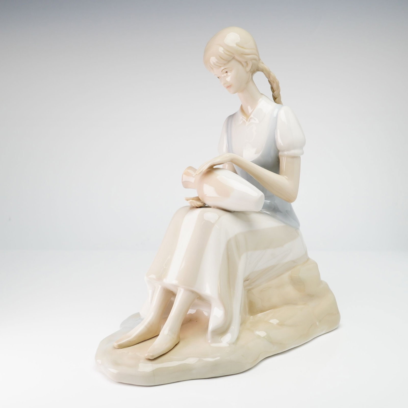 Italian Porcelain Figurine of a Seated Girl with Pottery: This glazed porcelain figurine depicts a seated young woman with blonde hair styled in two long braids. She is dressed in a pale blue bodice over a white short-sleeved blouse and a long, pale-colored