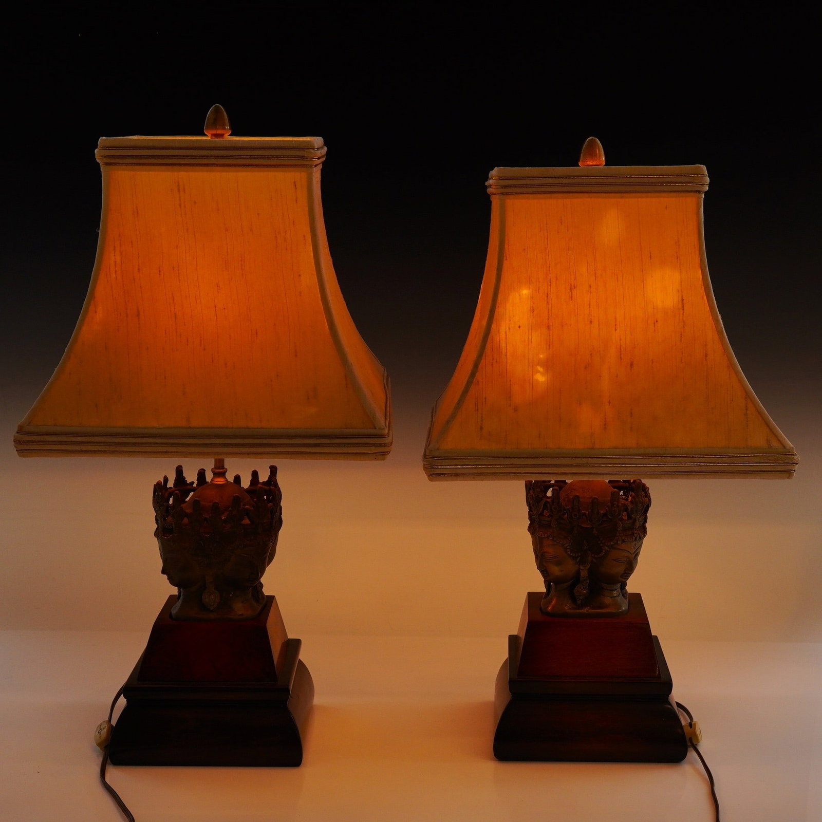 A Pair of Mid-Century Modern Chinoiserie Style Cast Metal and Wood Table Lamps: This pair of 2 lamps features central figural elements in the style of a four-faced Buddhist or Hindu deity, such as Brahma or Avalokiteshvara. Each face is depicted with serene features and elongated