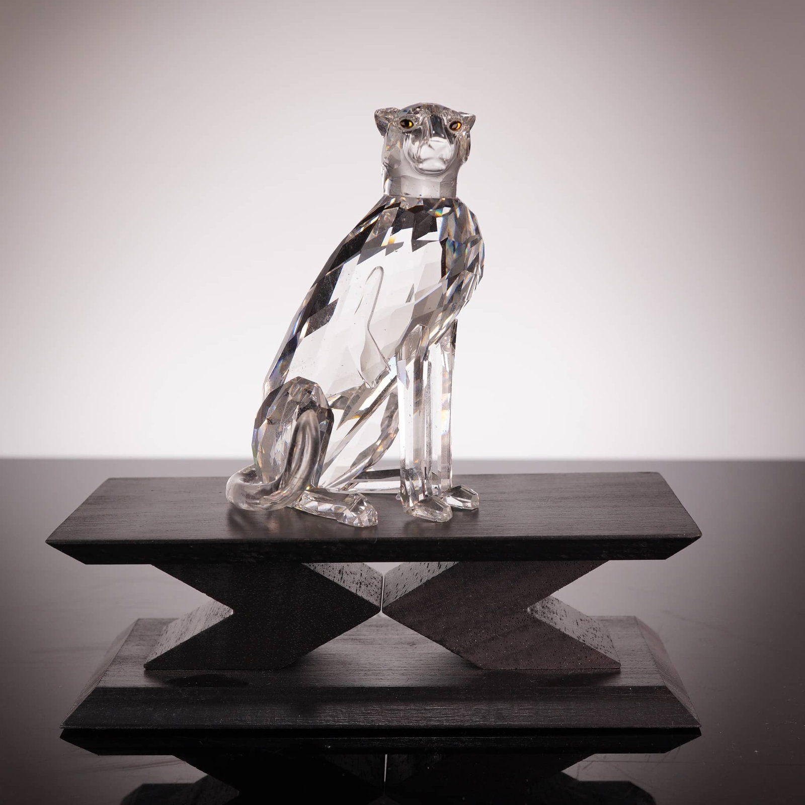 Swarovski Crystal Cheetah Figurine 183225 with Associated Black Stand: A Swarovski faceted clear crystal figurine of a seated cheetah, model 183225, featuring amber-toned eyes. The piece is designed in a contemporary style and is depicted with its tail in an upward posit