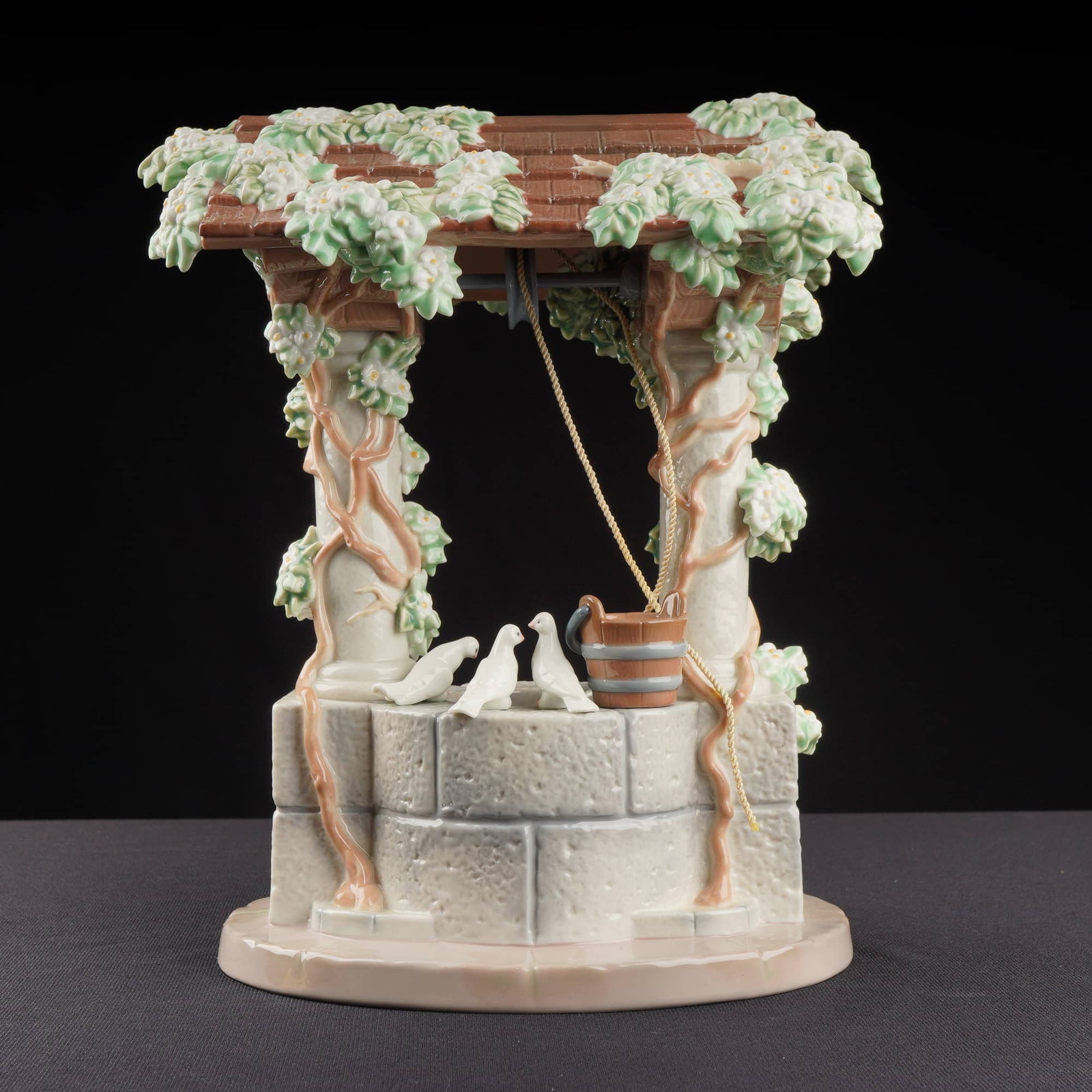 Lladro Porcelain Sculpture Snow Whites Wishing Well 01007558: The piece depicts a highly detailed Wishing Well structure featuring a stone block base, a tiled roof, and four supporting columns. The columns and roof are profusely decorated with applied ceramic el