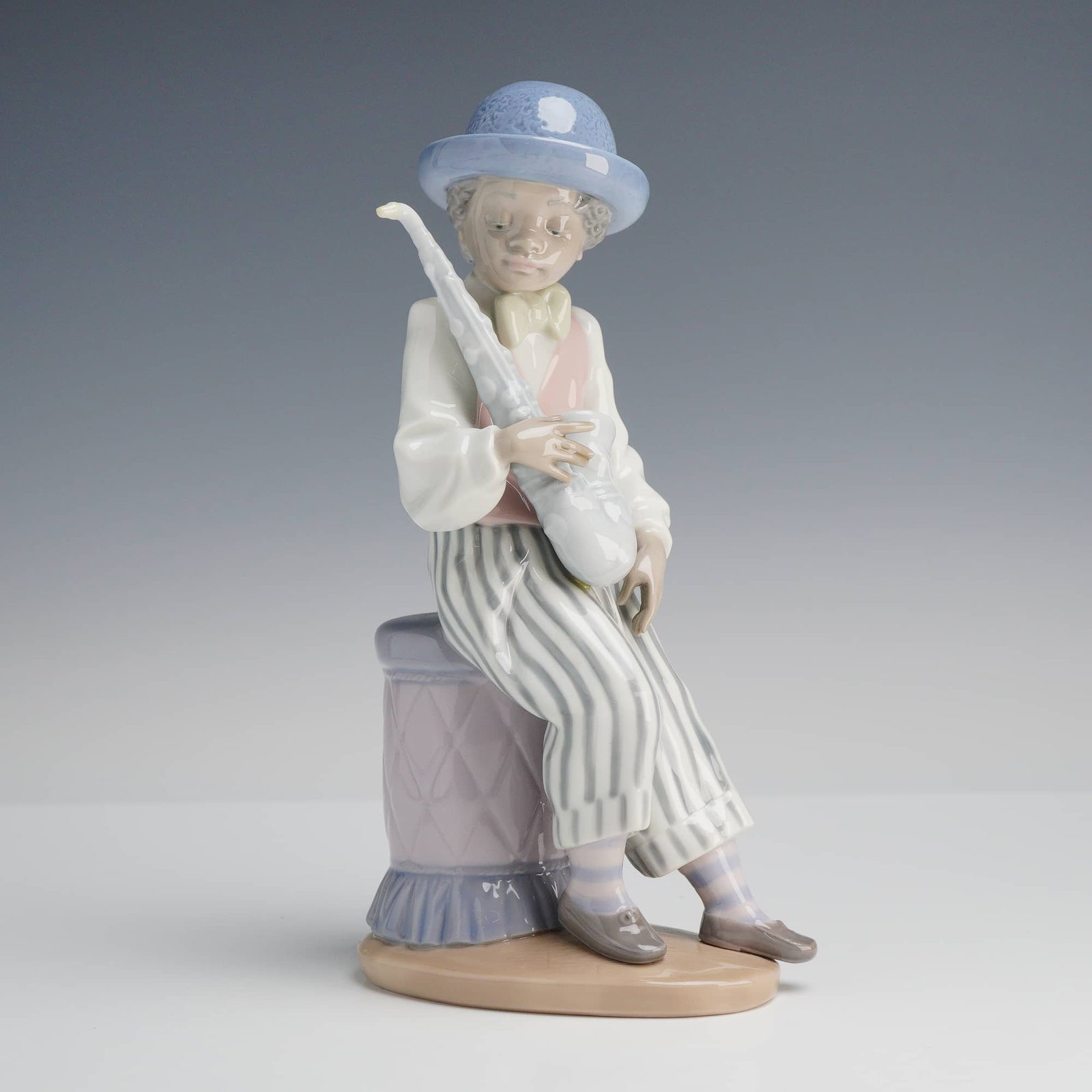 Lladró "Jazz Sax" Porcelain Figurine, Model 5133: This Lladró glazed porcelain figurine, titled "Jazz Sax" (model #5133), was designed by sculptor José Puche. The piece features a young boy musician seated on a quilted, ruffled stool, wearing a blu