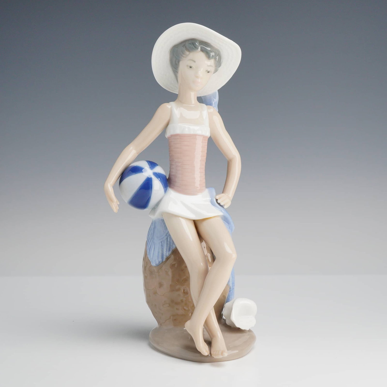 Lladró "Summer" Porcelain Figurine, Model 5219: This fine glazed porcelain Summer Lladró figurine, titled "Summer" (model #5219), was designed by Juan Huerta and originally issued in 1983. The piece depicts a young woman in a serene summer beach s