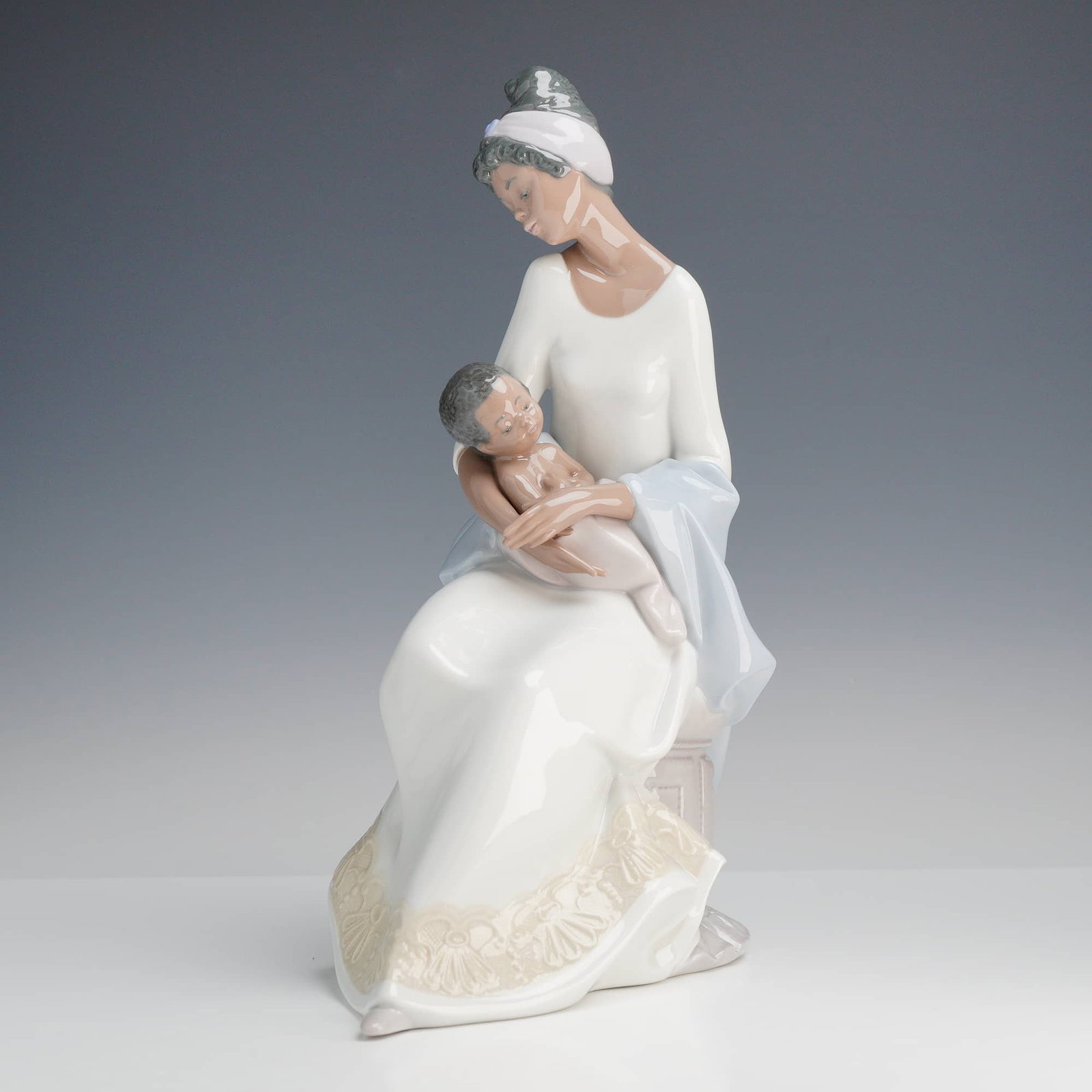 Lladró A Mother's Embrace Porcelain Figurine, Model 6851: A fine Lladró glazed porcelain figurine titled A Mother's Embrace. The piece depicts a poignant scene of a mother and child. The woman is seated on a low plinth, dressed in a flowing white gown with