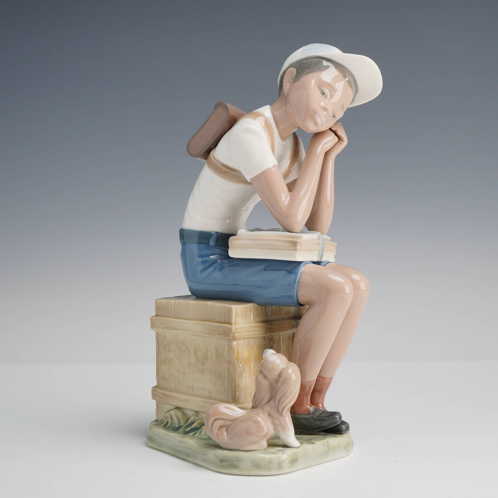 Lladró Exam Day Porcelain Figurine, Model 5250: A Lladró glazed porcelain figurine titled Exam Day, model number 5250, from the Black Legacy collection. The piece depicts a young schoolboy in a white baseball cap, white t-shirt, and blue shorts wi