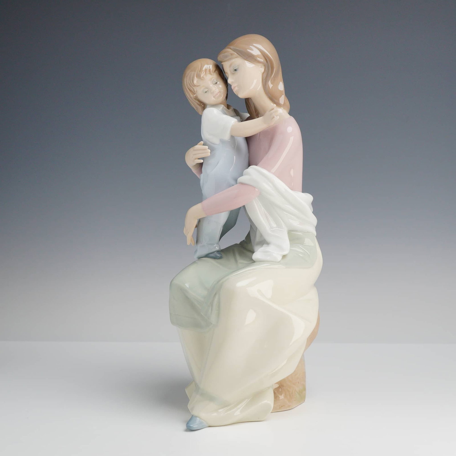 Lladró, A Mother's Love Figurine: This A Mother's Love Lladró figurine, model number 6634, is a high-gloss glazed porcelain sculpture depicting a tender scene of a mother seated on a rock while embracing her young child. The woman is