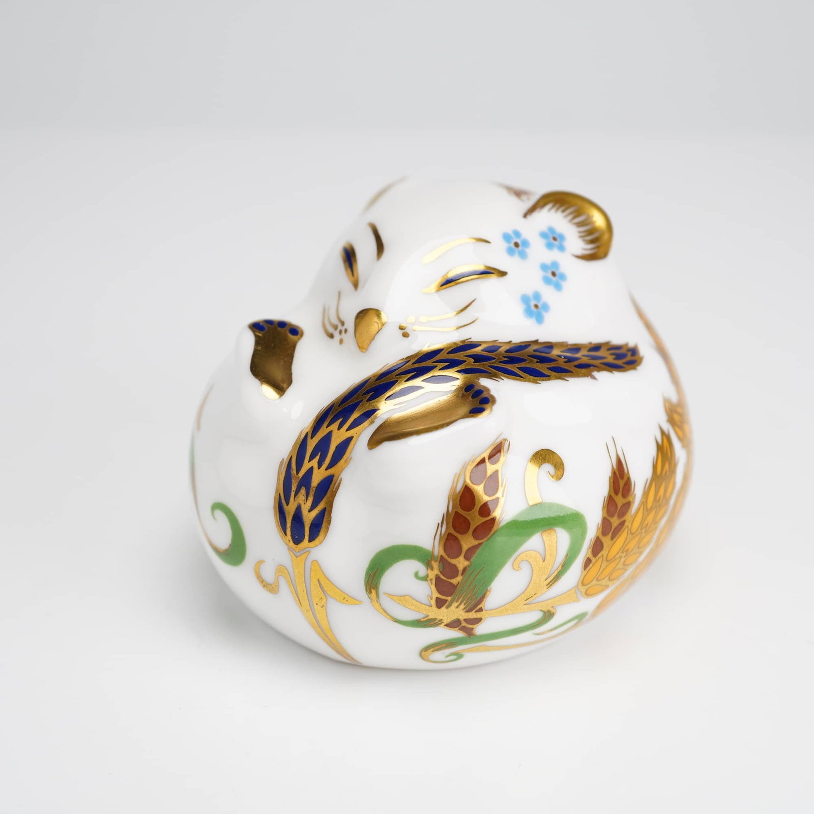 Royal Crown Derby Doormouse Paperweight: This Royal Crown Derby English bone china paperweight is modeled as a sleeping dormouse in a curled pose. The figurine is decorated with patterns of wheat ears, small blue flowers, and scrolling folia
