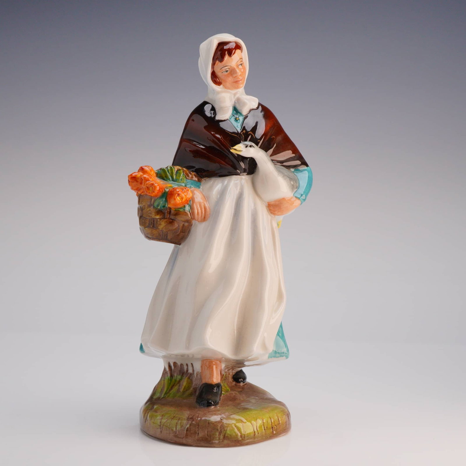 Royal Doulton Figurine Country Lass HN 1991: The figure depicts a standing woman dressed in a white bonnet, a dark brown shawl, a bright teal dress, and a white apron. She is shown holding a white goose under one arm and a brown basket filled wi