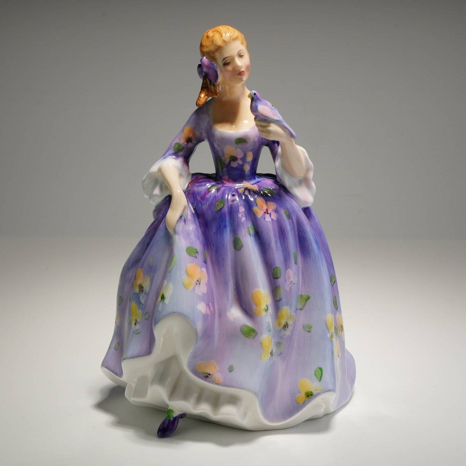 Royal Doulton Figurine Nicola HN2839: A bone china figurine manufactured Royal Doulton, titled Nicola, model HN3176. The figure depicts a woman in a purple, blue and light pink floral dress holding a small purple bird. The underside featu