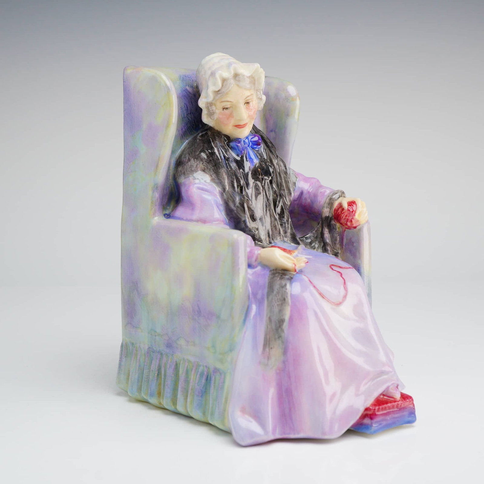 Royal Doulton Porcelain Figurine, Joan HN1422: A Royal Doulton glazed porcelain figurine titled Joan, model number HN1422. The piece depicts an elderly woman seated in a high-backed wing chair. The figure is dressed in a lavender gown with a black