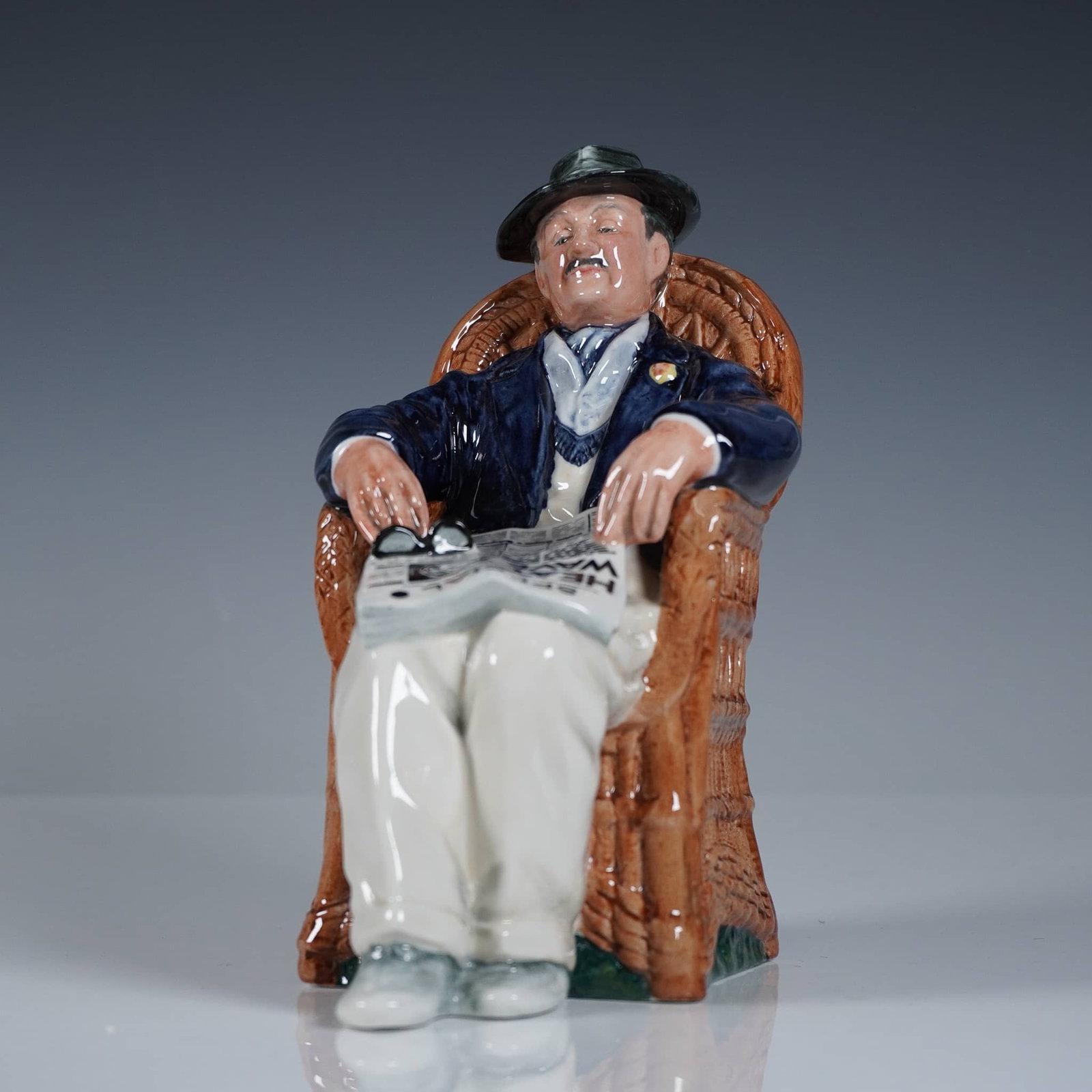 Royal Doulton Porcelain Figurine, Taking Things Easy HN2677: The hand-painted glazed figure depicts a seated elderly man in a wicker armchair, dressed in a dark jacket and light trousers, with a newspaper and eyeglasses resting on his lap. The subject's relaxed