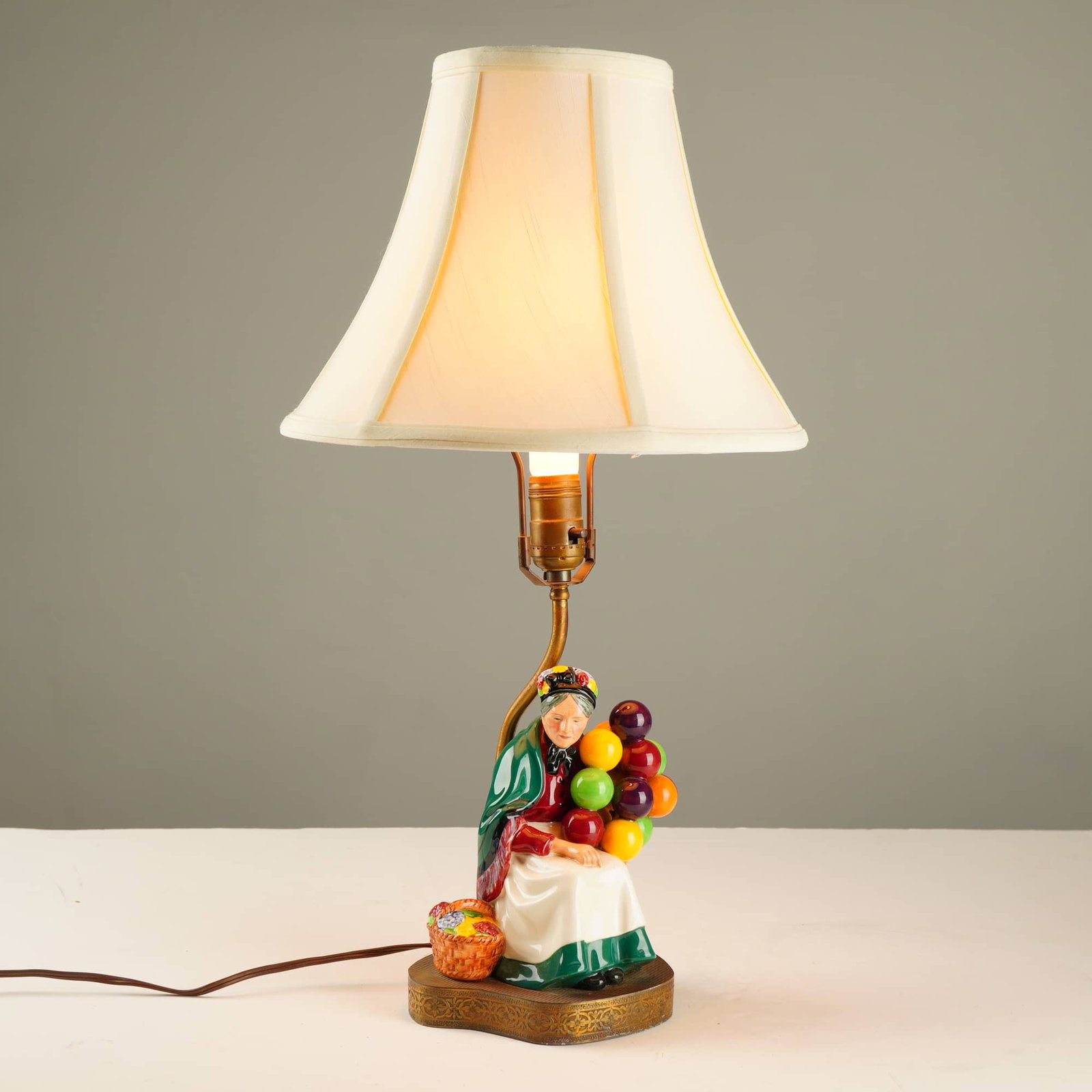 Royal Doulton The Old Balloon Seller Table Lamp Model HN 1315: This Royal Doulton The Old Balloon Seller table lamp, model HN 1315, was designed by Leslie Harradine. The piece features a hand-painted glazed ceramic figurine depicting an elderly woman seated with