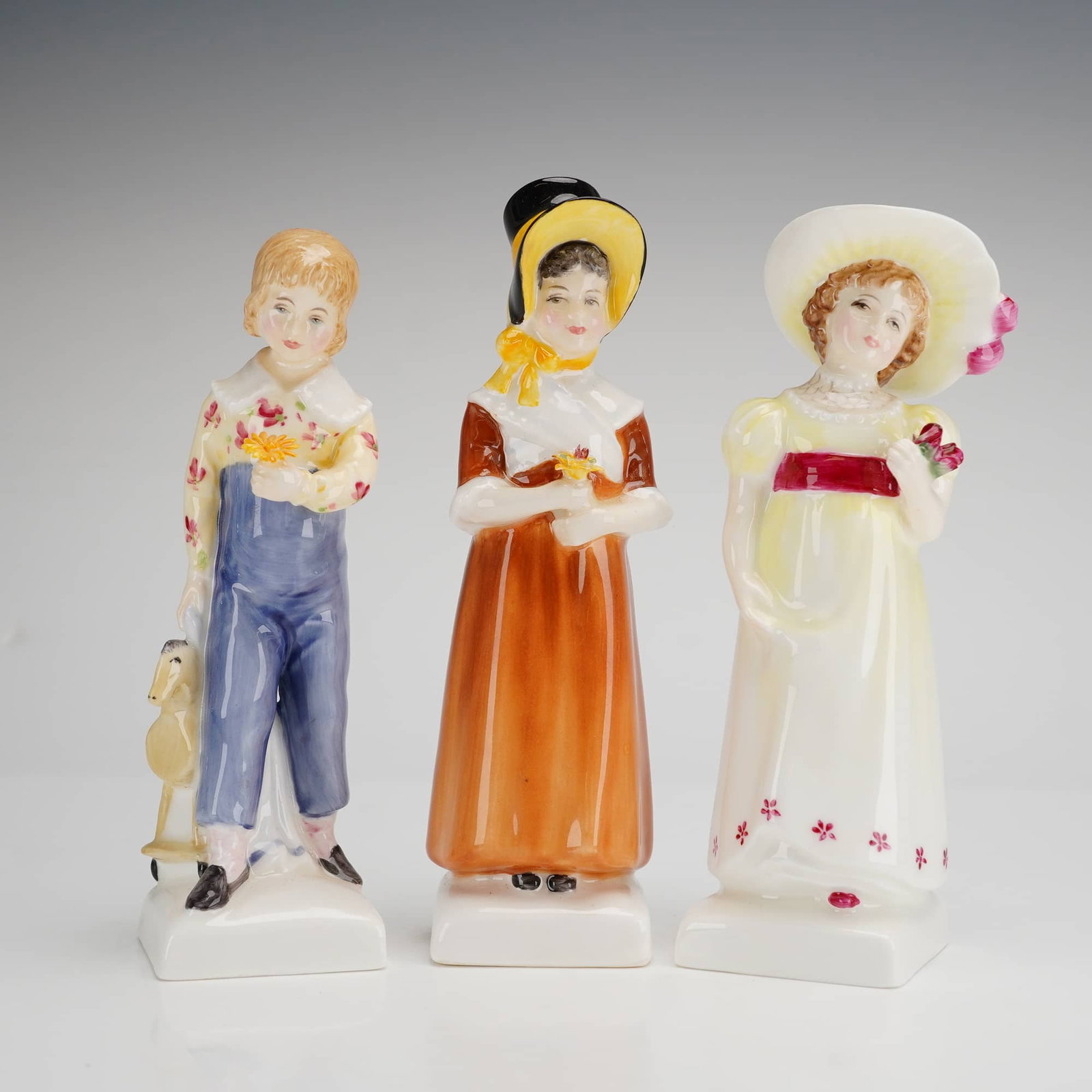 Three Royal Doulton Kate Greenaway Figurines Louise Tom Lori: This group of three Royal Doulton bone china figurines is from the Kate Greenaway Collection and includes the models Louise (HN2869), Tom (HN2864), and Lori (HN2801). Each figure is hand-painted and d