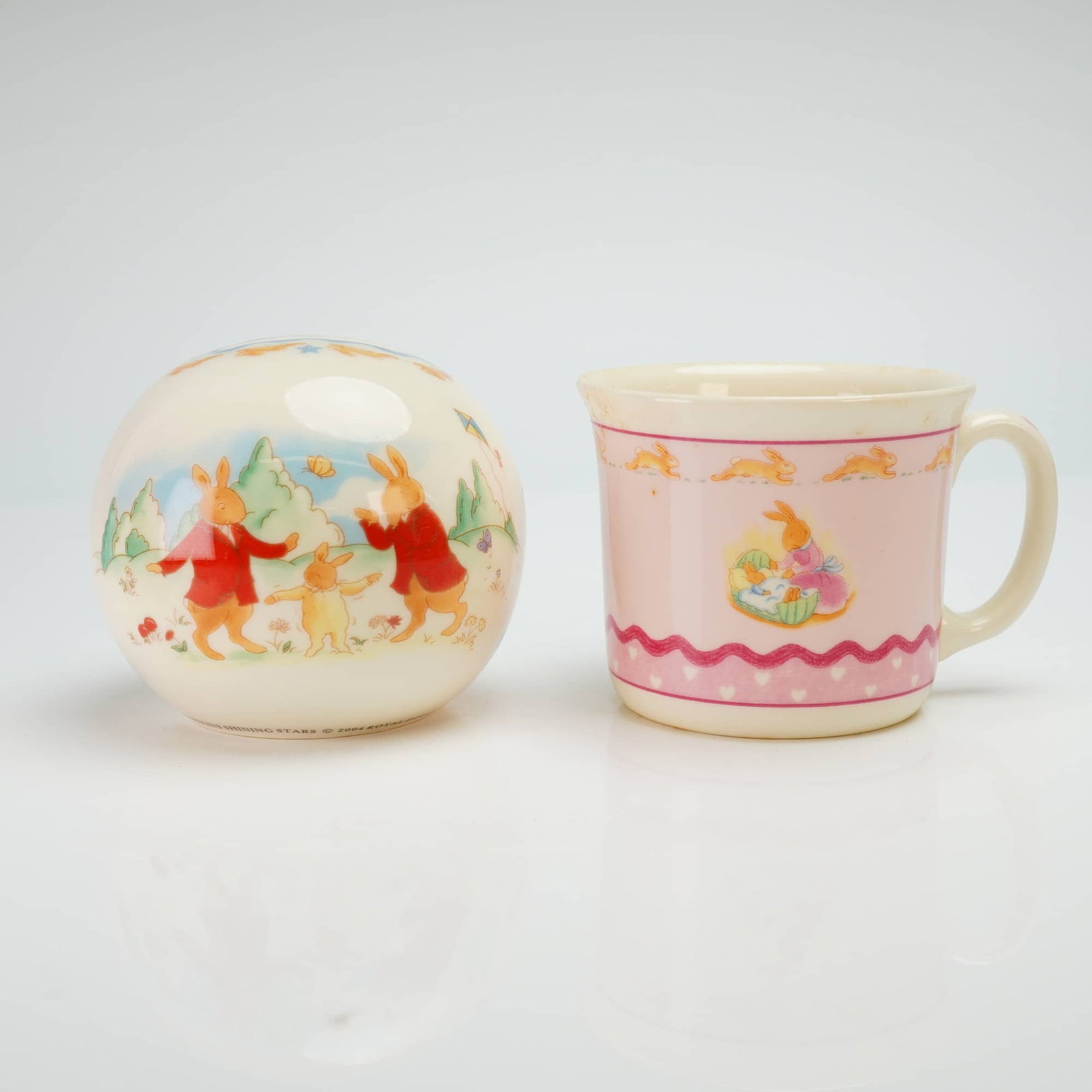 2pc Royal Doulton Bunnykins Bone China Money Ball and Mug: A 2pc Royal Doulton Bunnykins bone china set consisting of a money ball and a mug. The spherical money ball is decorated in the Shining Stars pattern, dated 2004, featuring transfer-printed scenes of