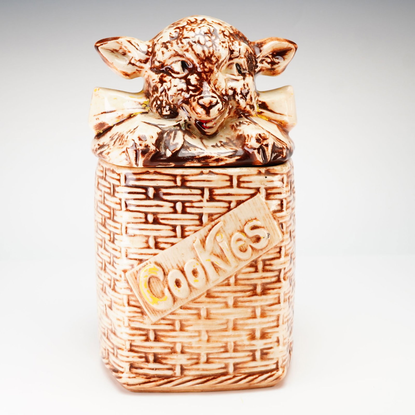 McCoy Lamb on Basketweave Cookie Jar: A mid-century McCoy Lamb on Basketweave cookie jar, produced circa 1950s. This ceramic vessel features a lid molded in the shape of a lamb's head and forepaws with a decorative bow. The rectangular ba