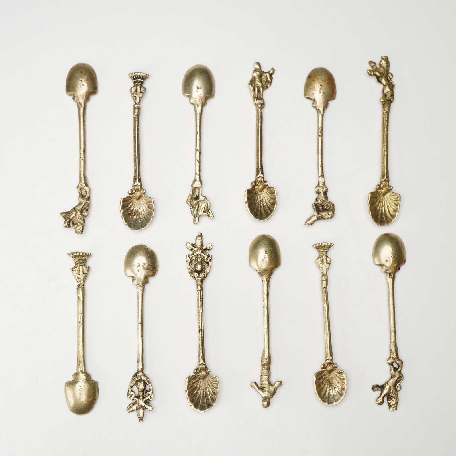 12pc Salt Spoon Set from Italy: A set of twelve salt spoons from Italy manufactured from gold-tone metal, likely brass. Each spoon features a unique decorative finial or handle motif. These motifs include the Capitoline Wolf, herald