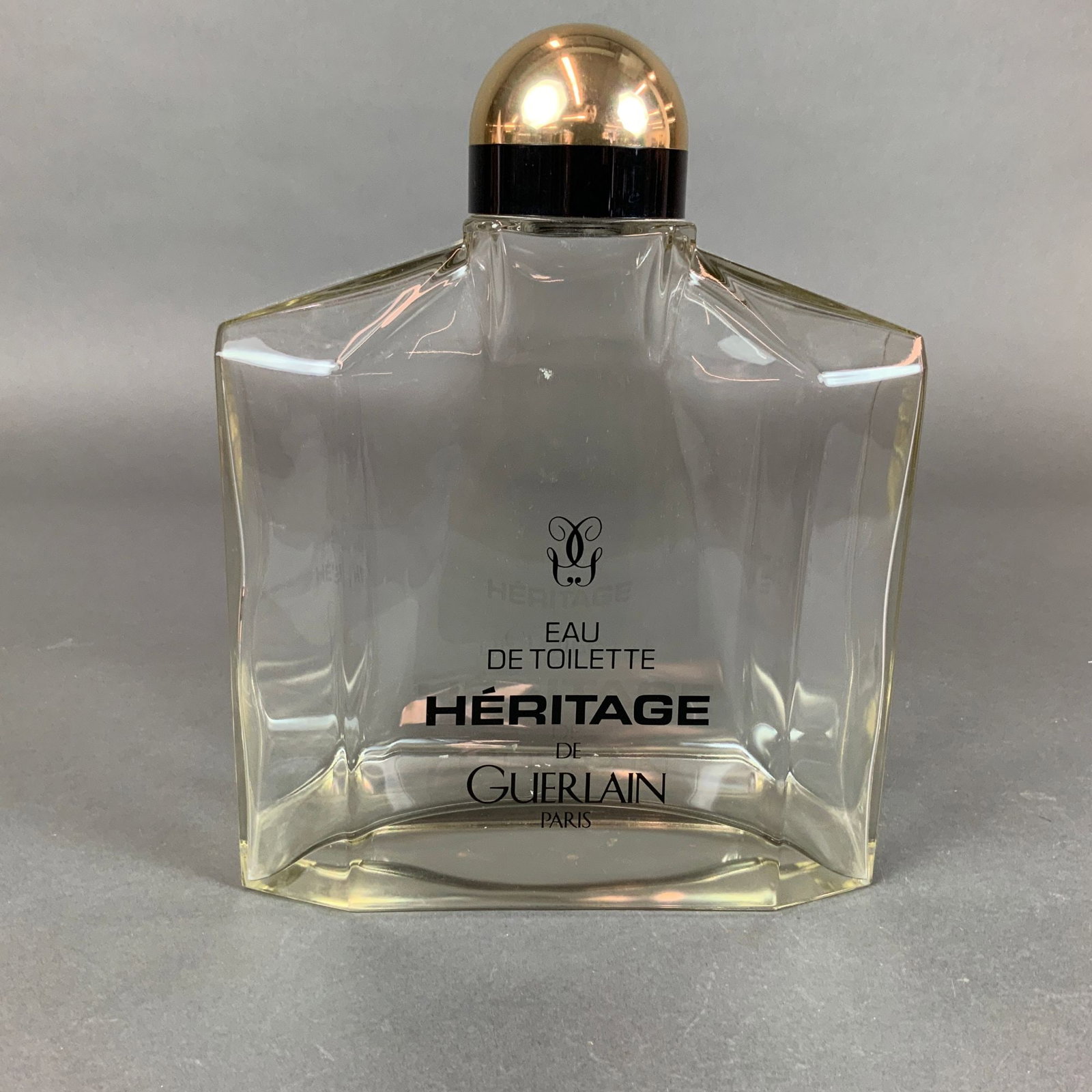 Heritage de Guerlain Large Eau de Toilette Glass Bottle: A large clear glass perfume bottle for Heritage de Guerlain. The bottle features a broad, faceted rectangular form with a black plastic collar and a gold-toned domed cap. The front of the bottle is pr
