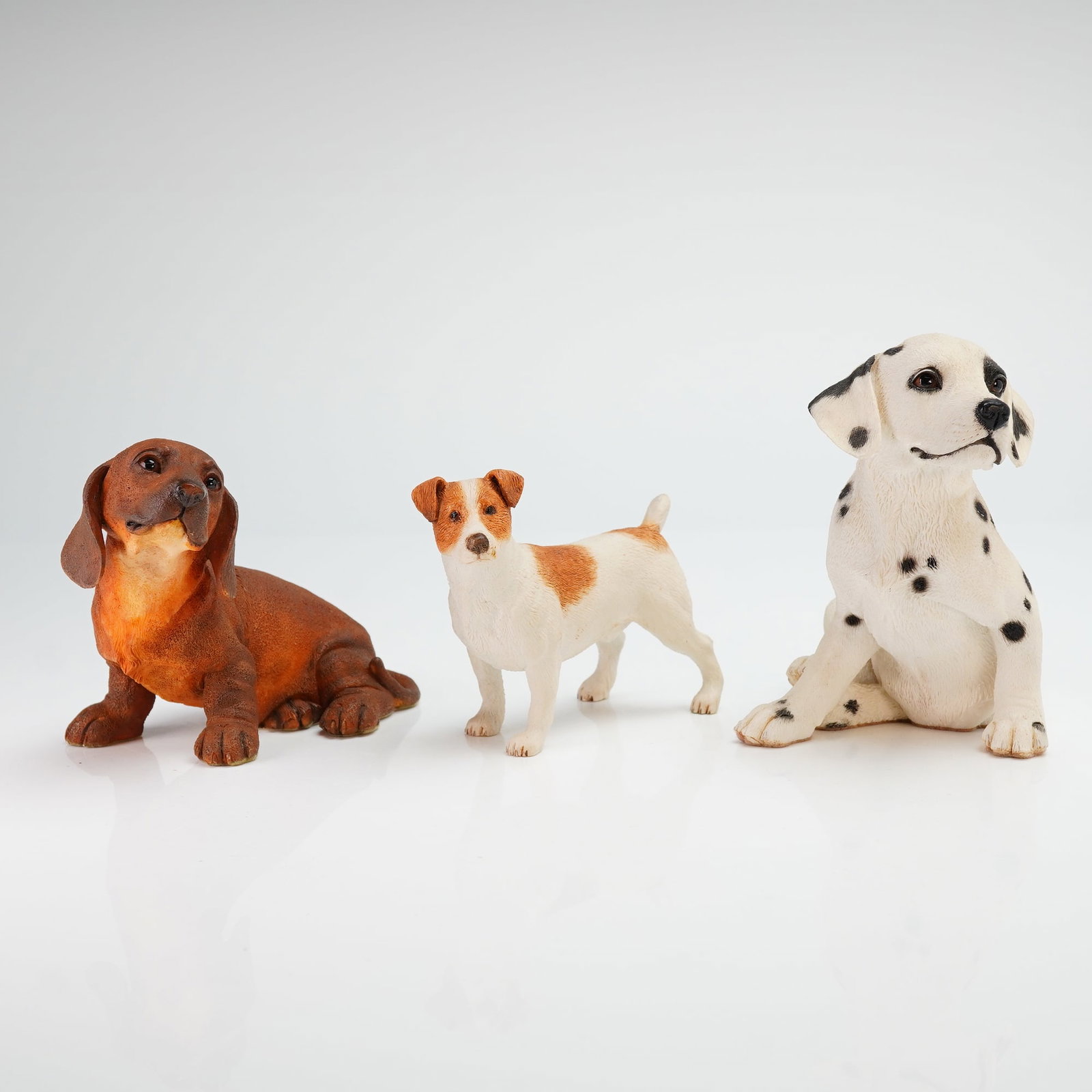Three Country Artists England Dog Figurines: This set includes a tan Dachshund puppy, a Jack Russell Terrier, and a Dalmatian puppy. Each figure is constructed from cast resin with hand-painted naturalistic coloring and detailed fur textures. Ma