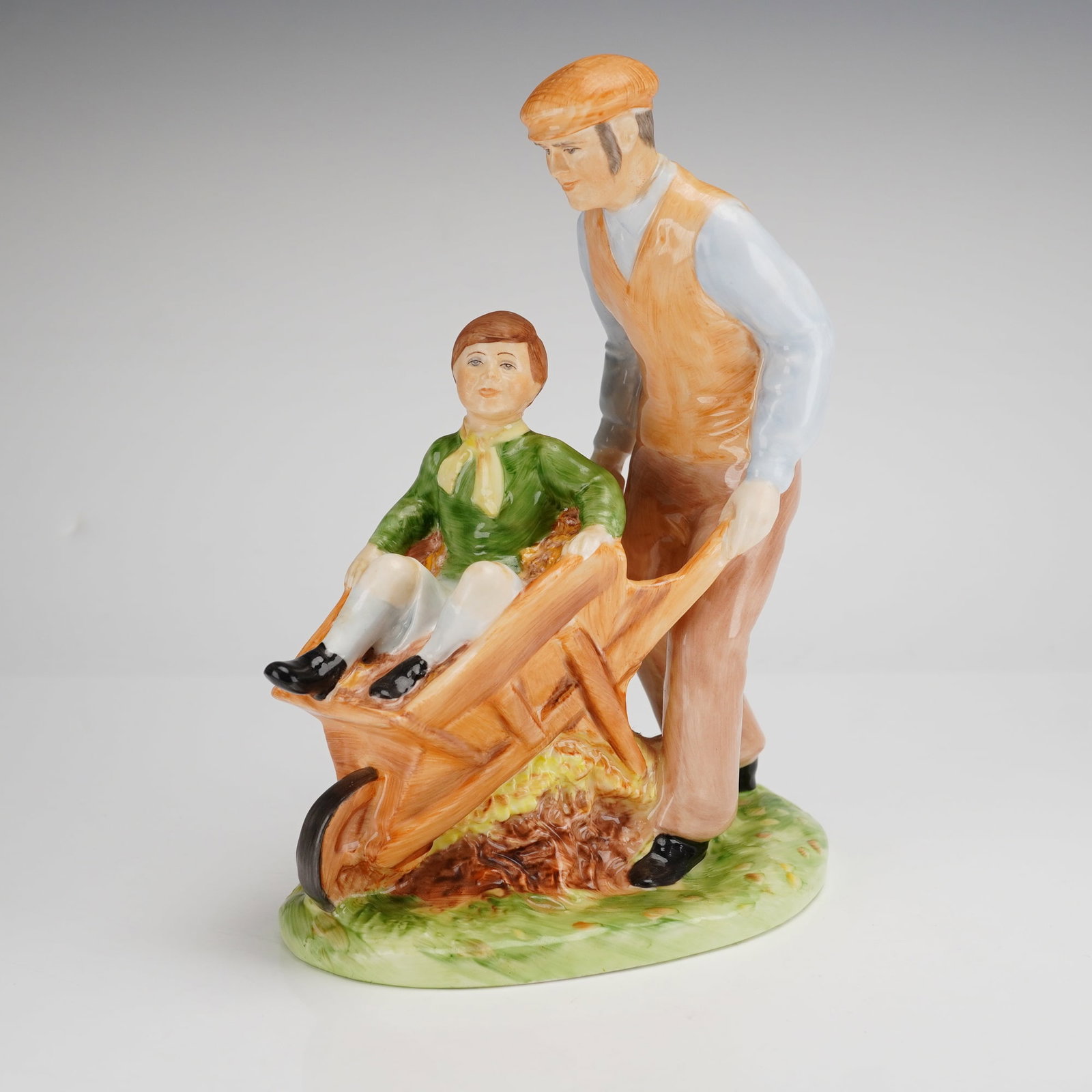 Coalport Bone China Figurine, Autumn Leaves: Figure is of a modeled man in a flat cap and orange vest pushing a young boy in a wooden-style wheelbarrow over a pile of fallen leaves. The piece is hand-painted in a naturalistic polychrome palette