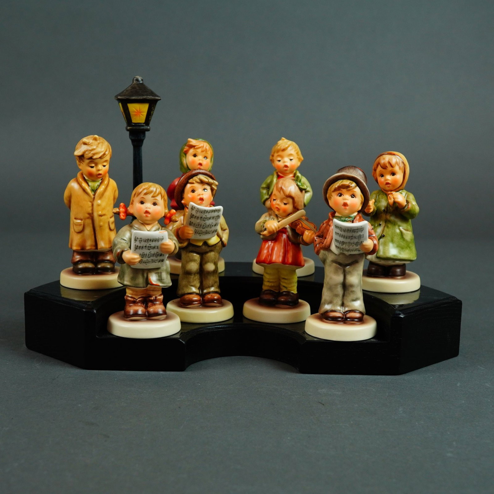Goebel M.I. Hummel Kinder Choir 8-Piece Figurine Set with Lamppost Display Stand: A comprehensive collection of eight hand-painted porcelain figurines from the Goebel M.I. Hummel Club series, collectively known as the Kinder Choir. This set features a group of children in tradition