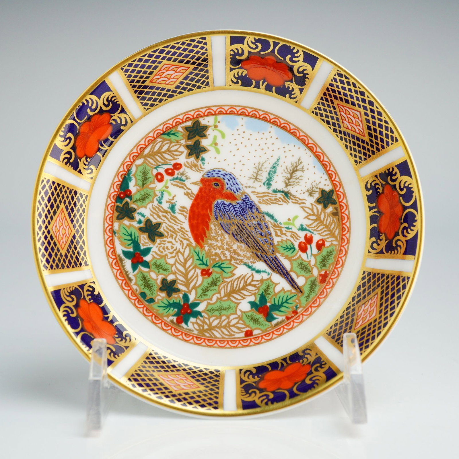 Royal Crown Derby Bone China Tray, Old Imari Christmas Robin: This circular piece features a central medallion depicting a robin perched among holly leaves and red berries within a stylized winter landscape. The decoration is executed in the traditional Imari pa