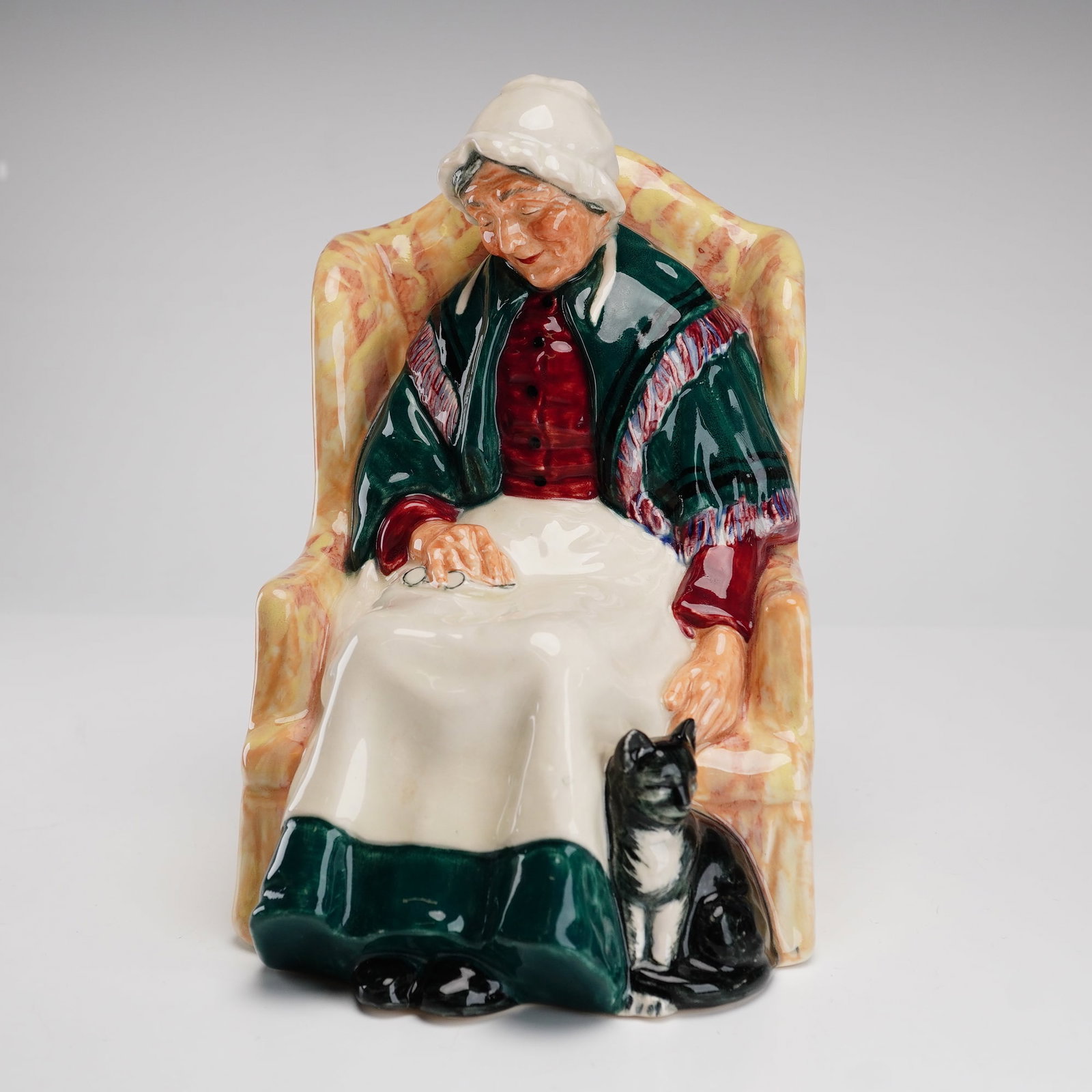 Royal Doulton Figurine, Forty Winks HN1974: The piece depicts a charming domestic scene of an elderly woman asleep in a high-backed wing chair, accompanied by a black cat at her feet. She is dressed in a white bonnet, a green fringed shawl over