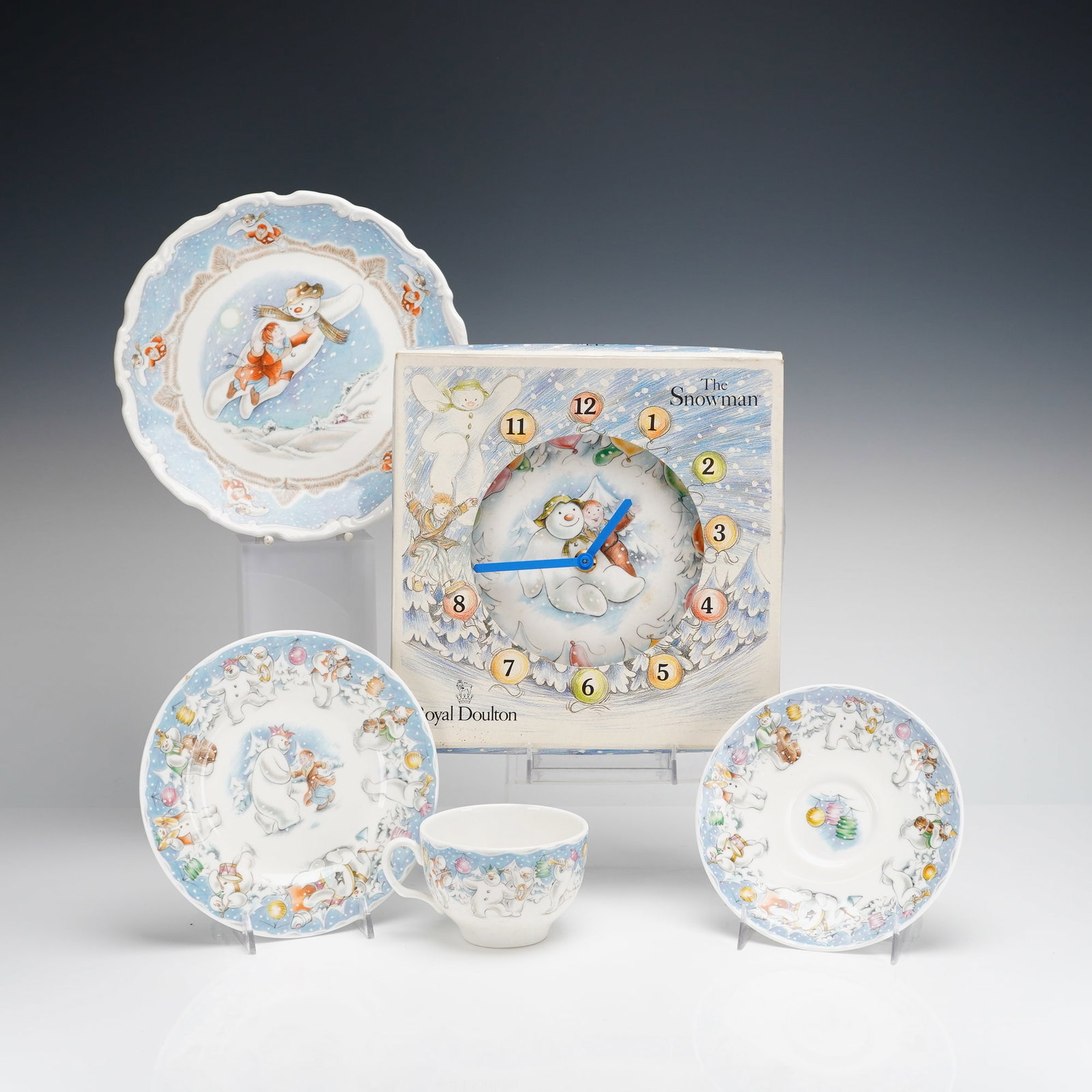 5pc Royal Doulton The Snowman Gift Collection Clock, Cup, Saucer, & Plates: A 5-piece set of Royal Doulton fine bone china from The Snowman Gift Collection, featuring illustrations based on the work of Raymond Briggs. The group includes a wall clock in its original illustrate