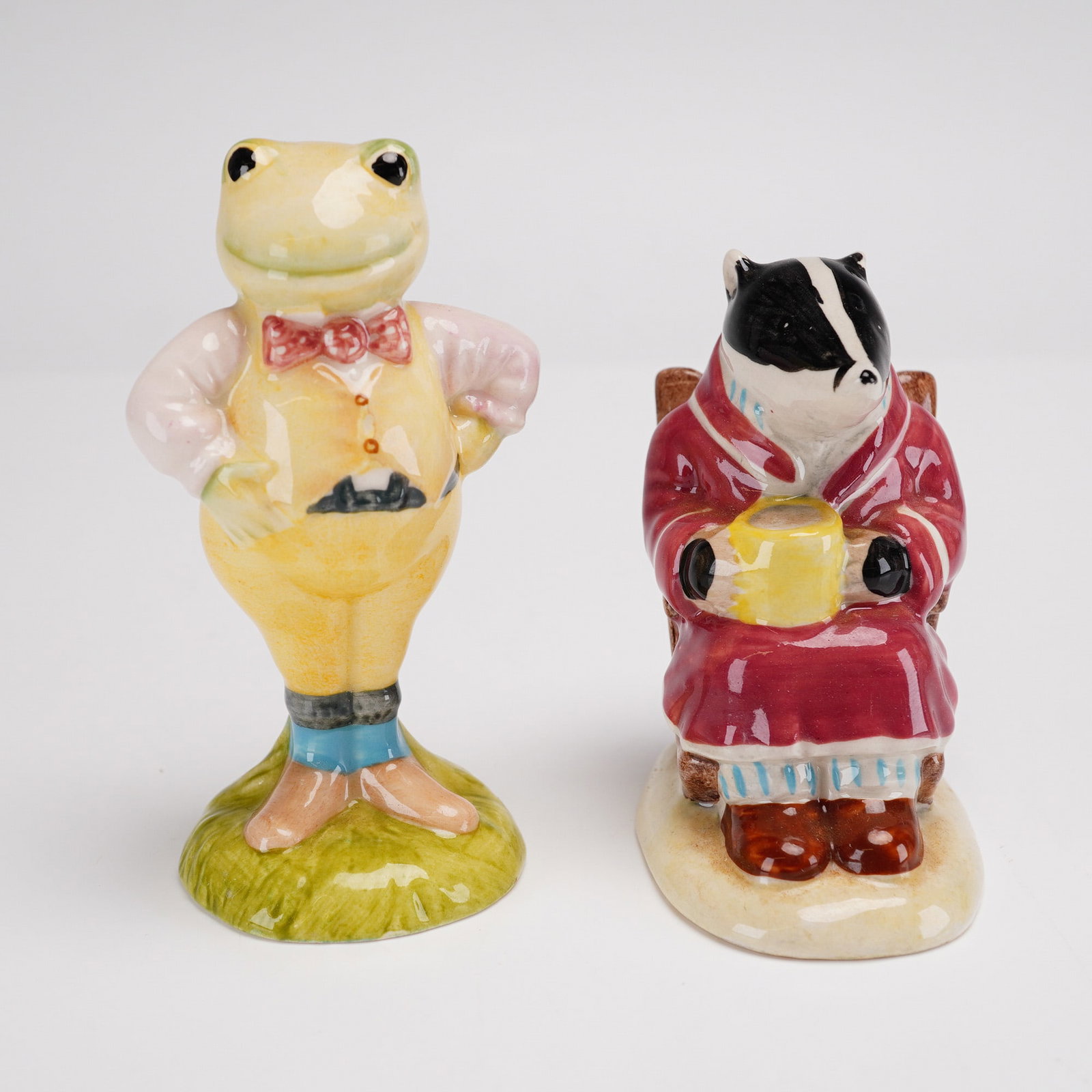2pc Royal Albert Figurines, Toad (AW1) and Badger (AW2): This pair of fine glazed porcelain figurines was produced by Royal Albert in England as part of The Wind in the Willows series. The set includes Toad (model AW1), depicted in a yellow waistcoat and re