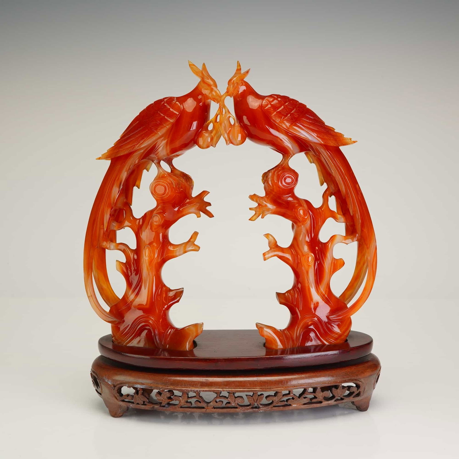 Japanese Orange Jade Birds of Paradise Figurine with Wooden Stand: The stone displays a vibrant reddish-orange color with characteristic translucent white and cream banding. The sculpture features two long-tailed paradise birds perched atop gnarled, openwork branches