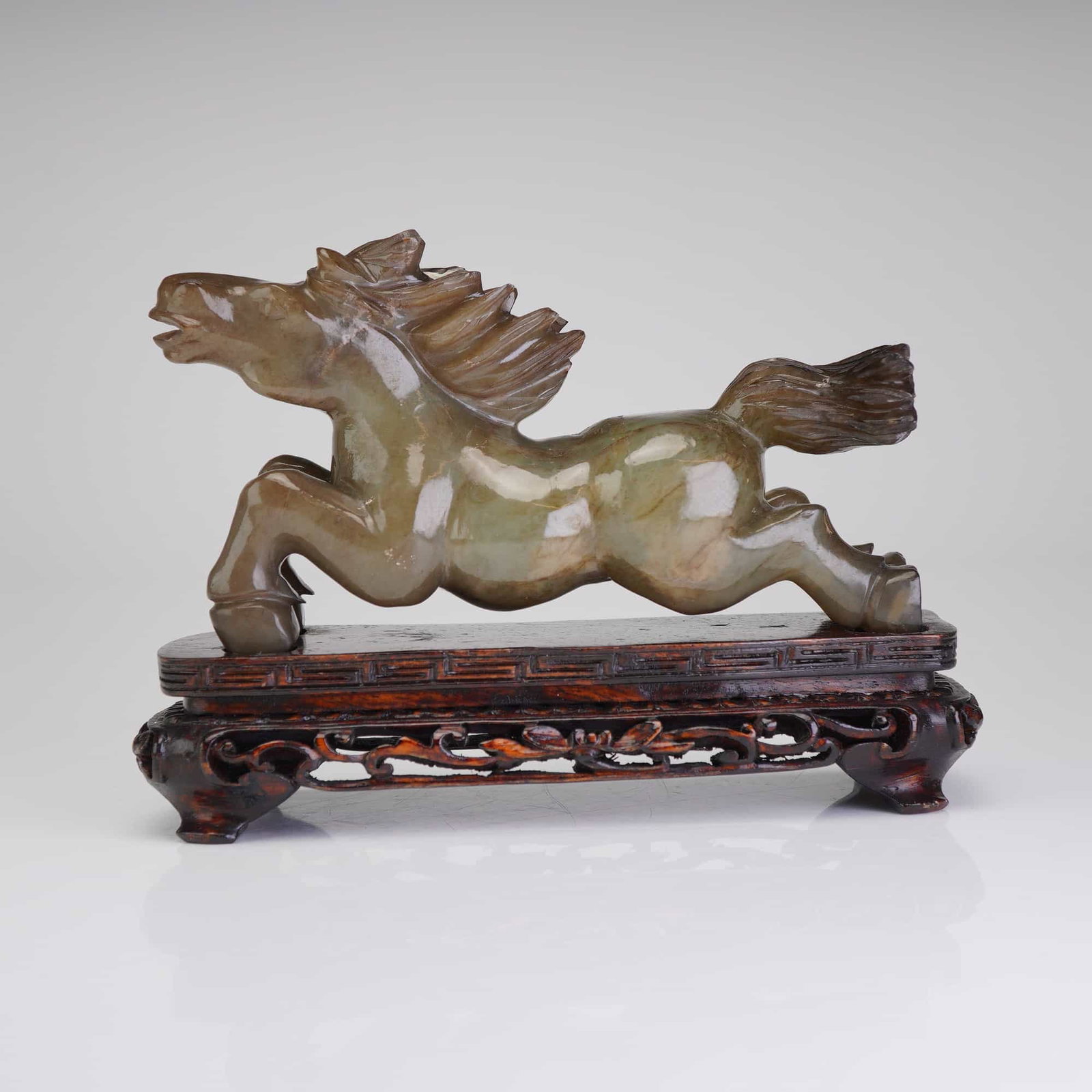 Chinese Carved Jade Horse Figurine on Wooden Stand: The stone exhibits a variegated palette ranging from olive and celadon green to deep russet and earthy brown. The horse is depicted in a dynamic galloping pose, with its musculature rendered in soft,