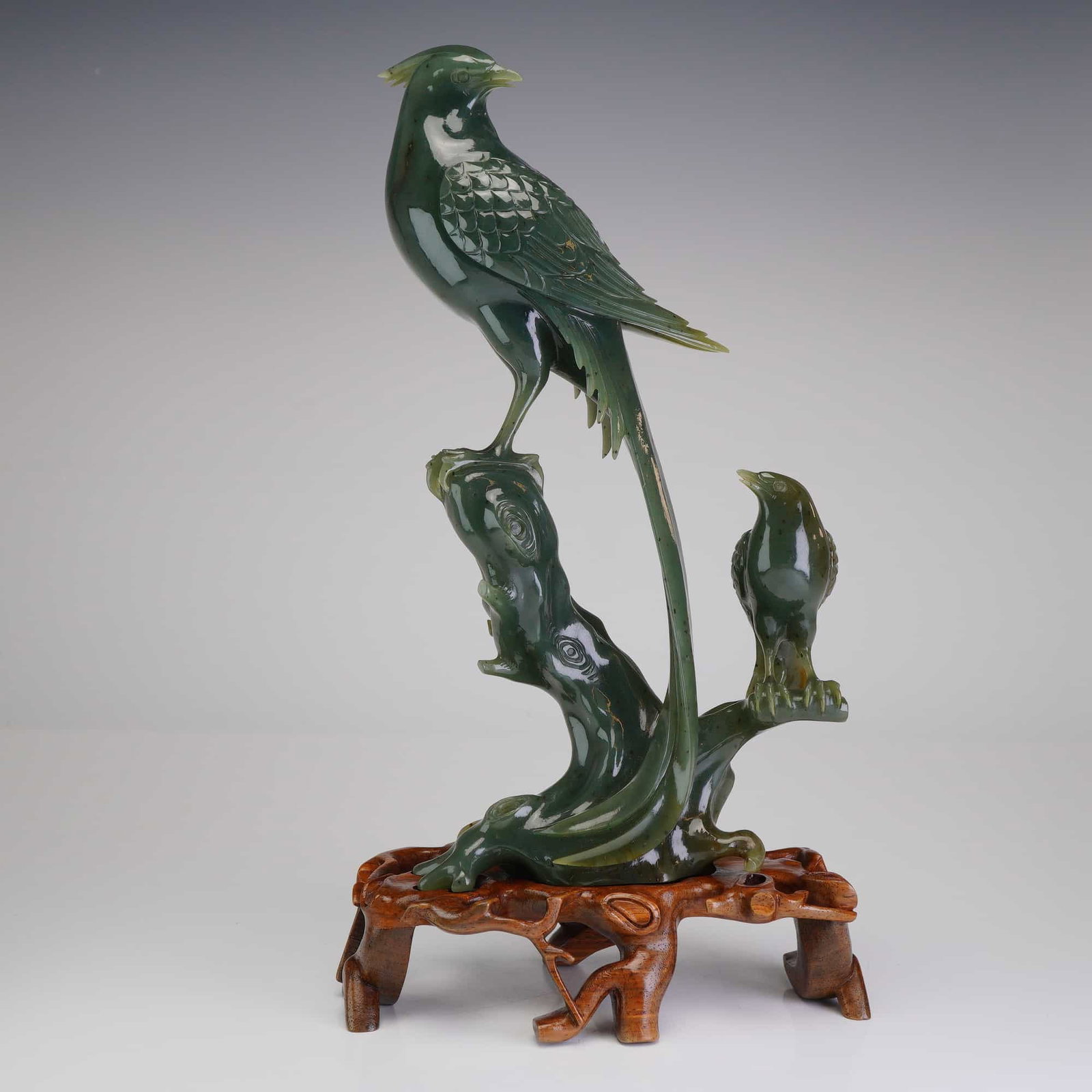 Chinese Spinach Jade Bird Figurine with Wooden Stand: This stone carved bird figurine features a large crested bird, such as a pheasant or paradise flycatcher, with detailed incised feathering and a long, sweeping tail. The primary bird is perched atop a